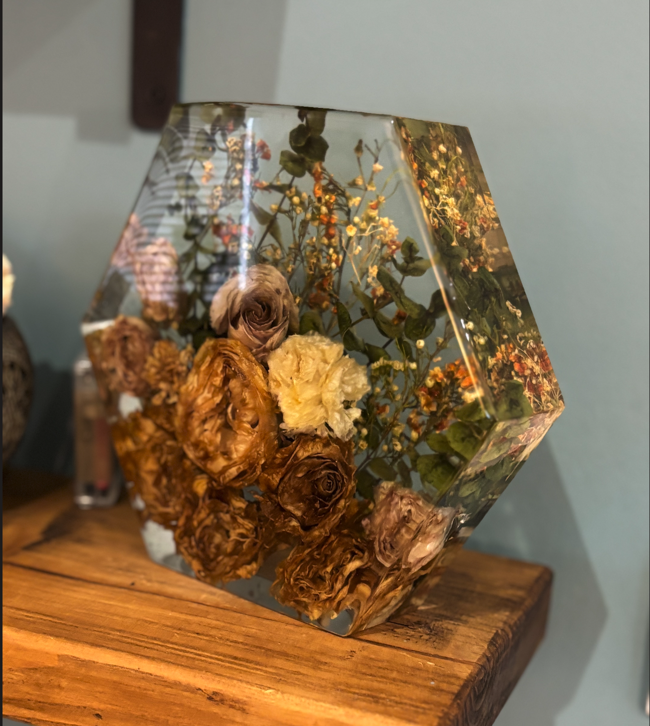 A clear, geometric-shaped glass vase with dried roses, foliage, and other preserved flowers, sitting on a wooden surface.