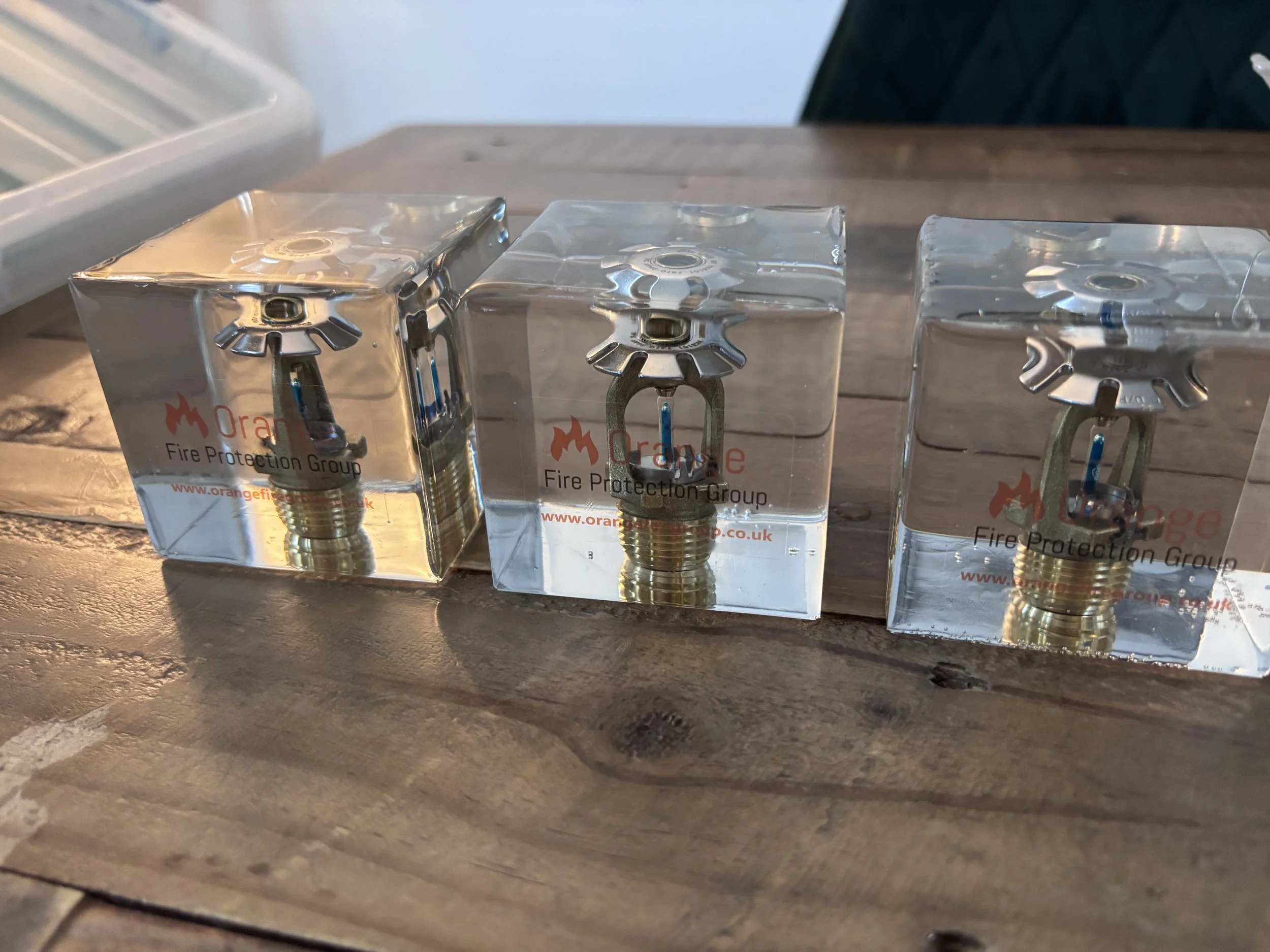 Three fire sprinkler heads encased in clear acrylic blocks with the 'Orange Fire Protection Group' logo visible on the front, placed on a wooden surface.