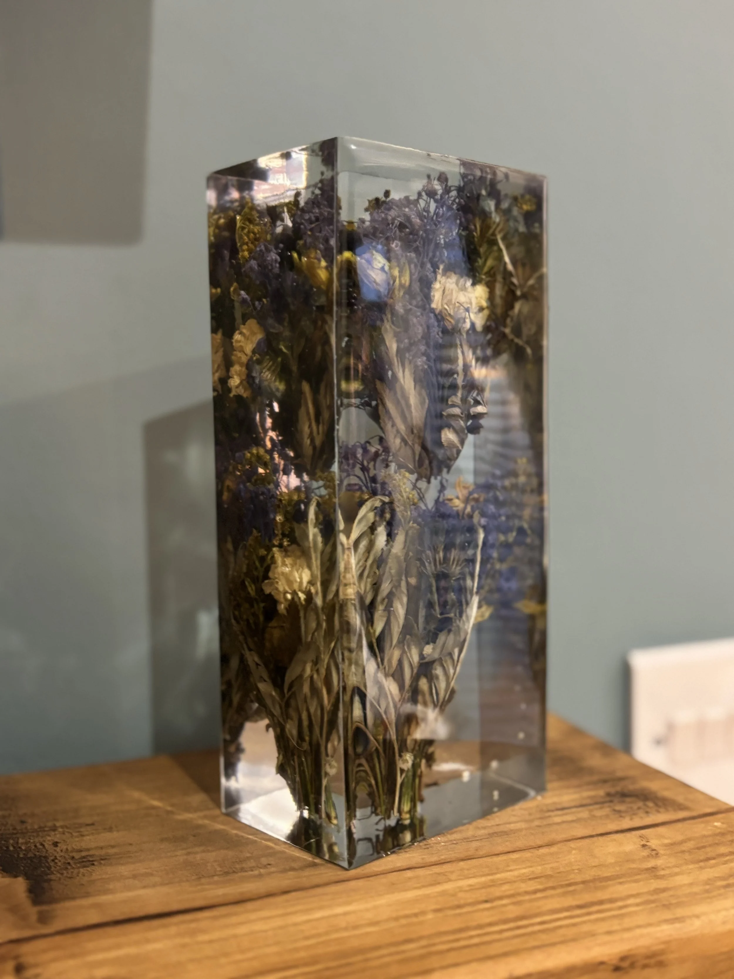 A clear rectangular resin block filled with dried purple and yellow flowers, placed on a wooden surface.