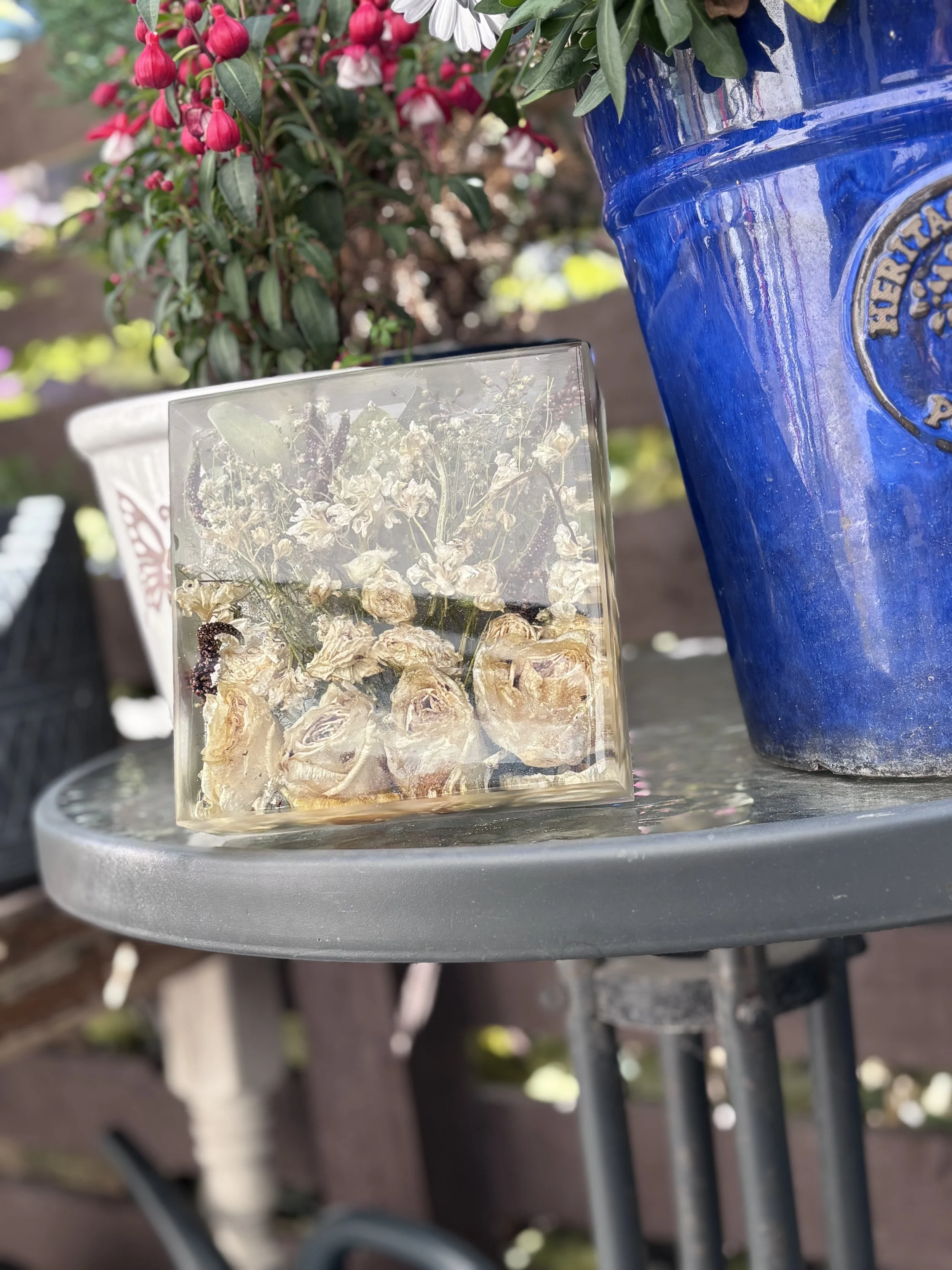 A small glass cube containing dried flowers is placed on a round metal table. To the right, there is a large blue ceramic planter with some embossed lettering. In the background, there are various potted plants with pink flowers and green leaves.