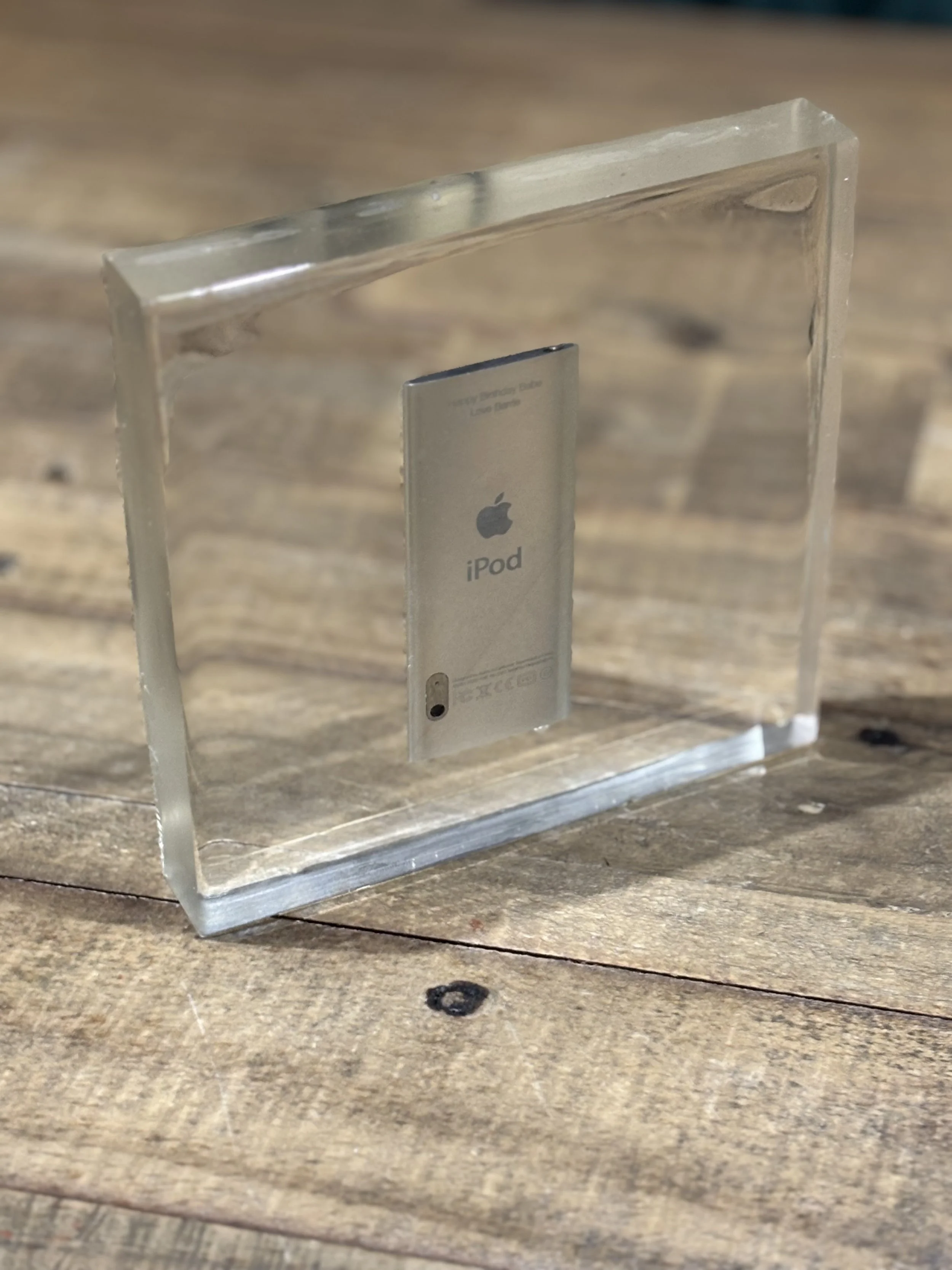 A clear acrylic display case holding a silver iPod, with the back side visible, placed on a wooden surface.
