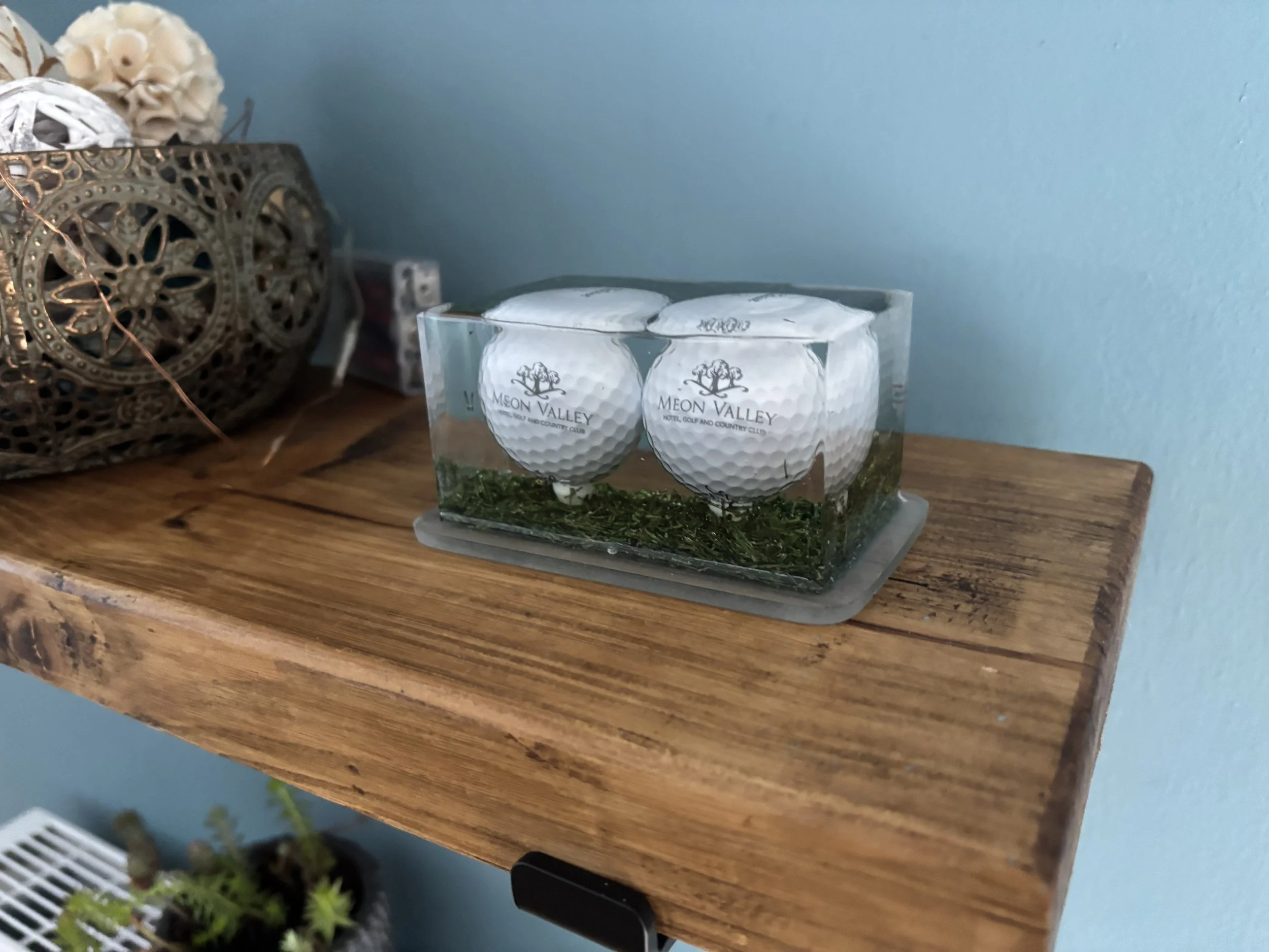 Decorative table with a set of two golf balls in a glass container labeled Meon Valley, resting on a wooden shelf with a decorative metal basket and flowers in the background.