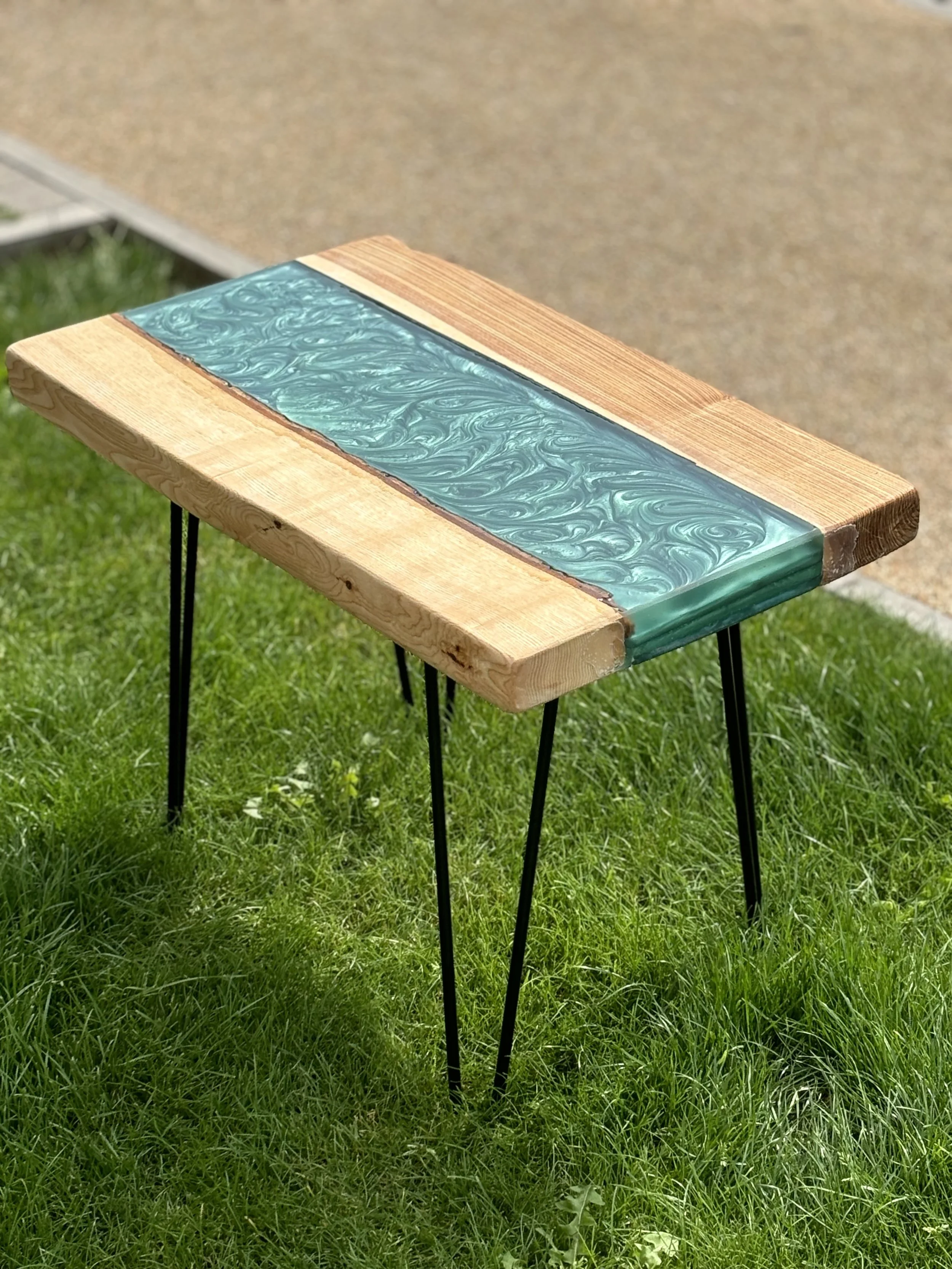 A small table with a wooden top and a decorative resin inlay sits on a grassy area outdoors.
