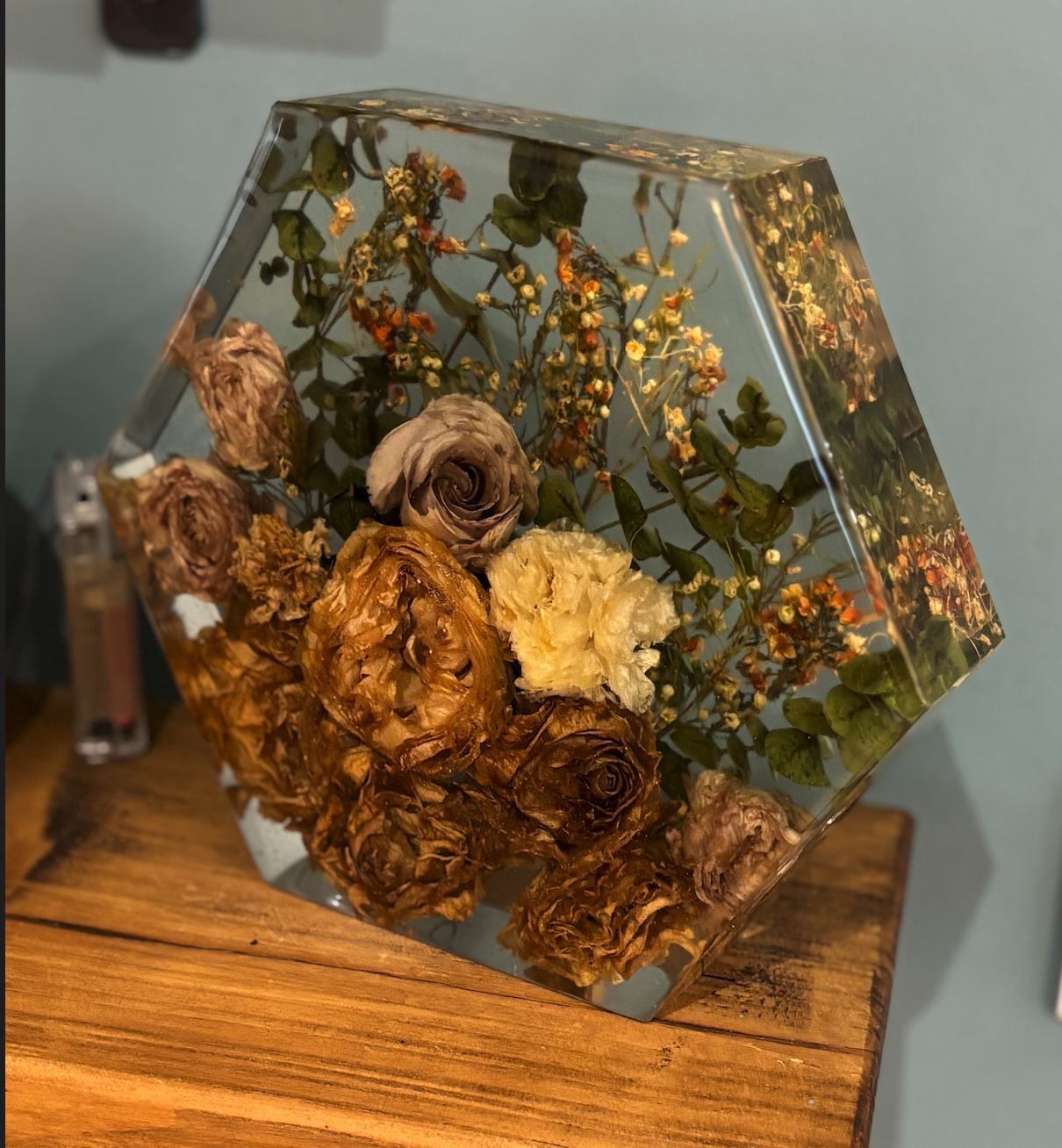 A square glass container with dried roses and small flowers inside, placed on a wooden surface.