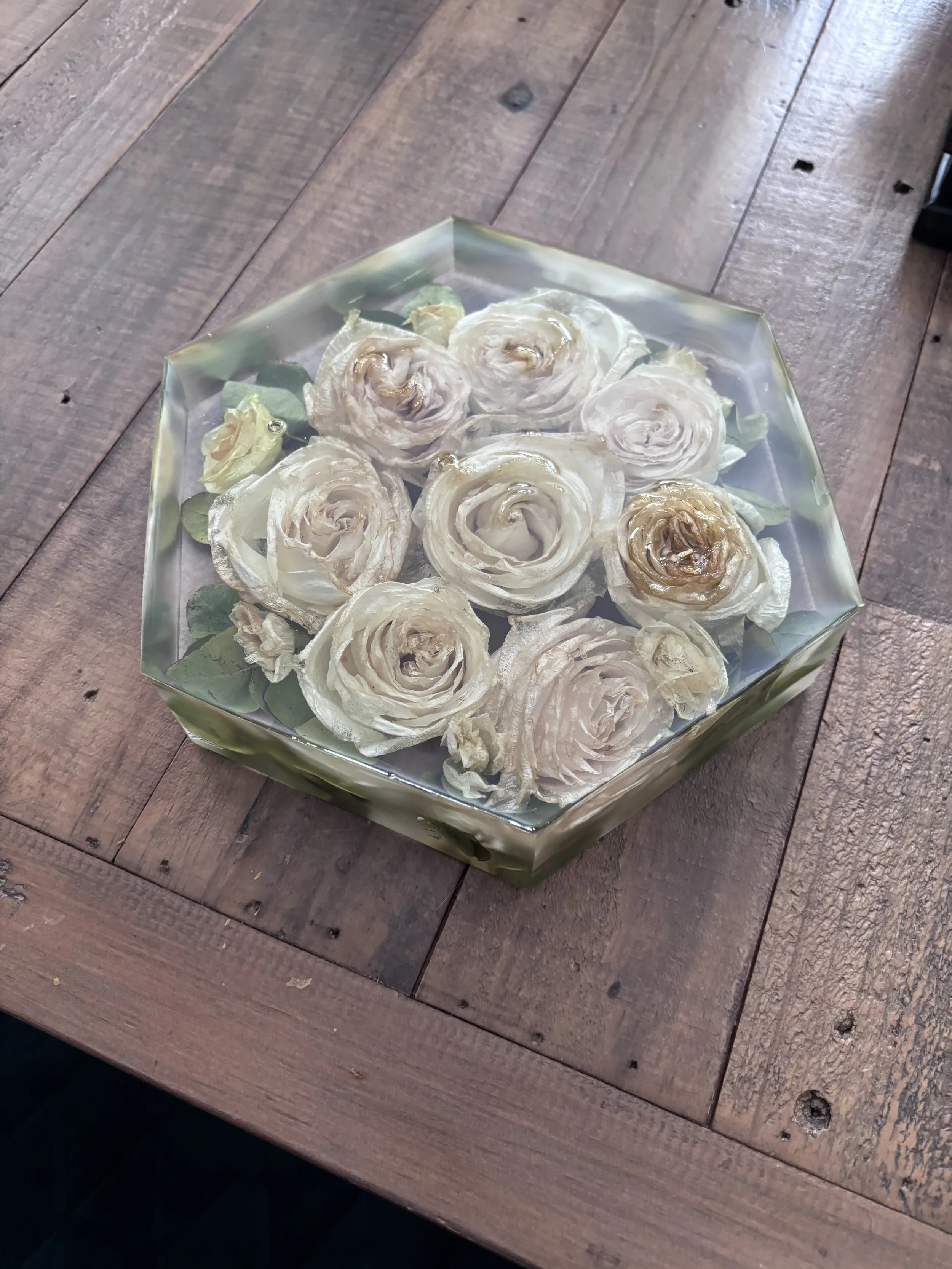 A square glass container with a floral design, containing dried white and beige roses with green leaves, sitting on a wooden surface.