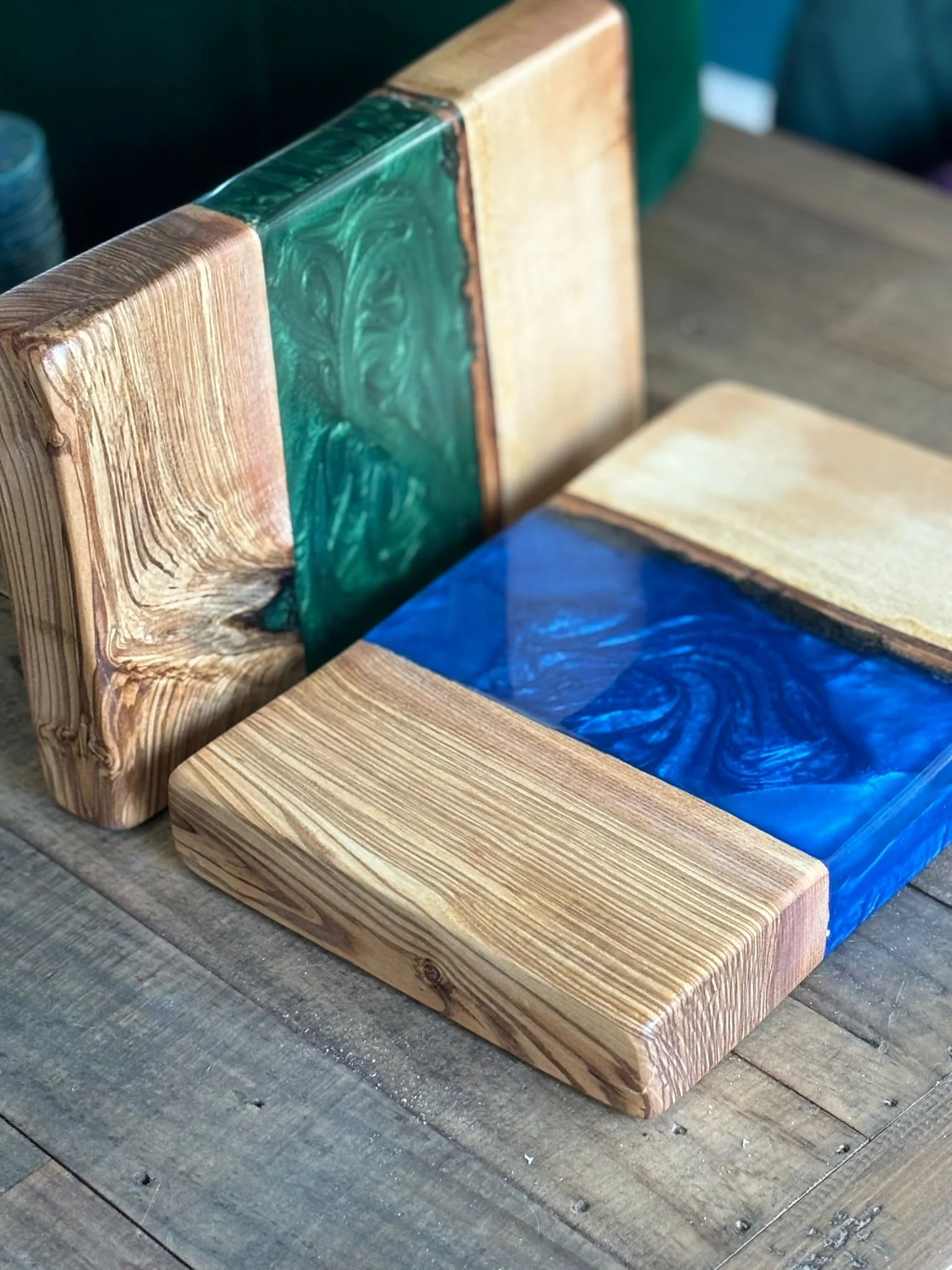 Wooden cutting boards with resin inlays, one with green resin, the other with blue, placed on a wooden surface.