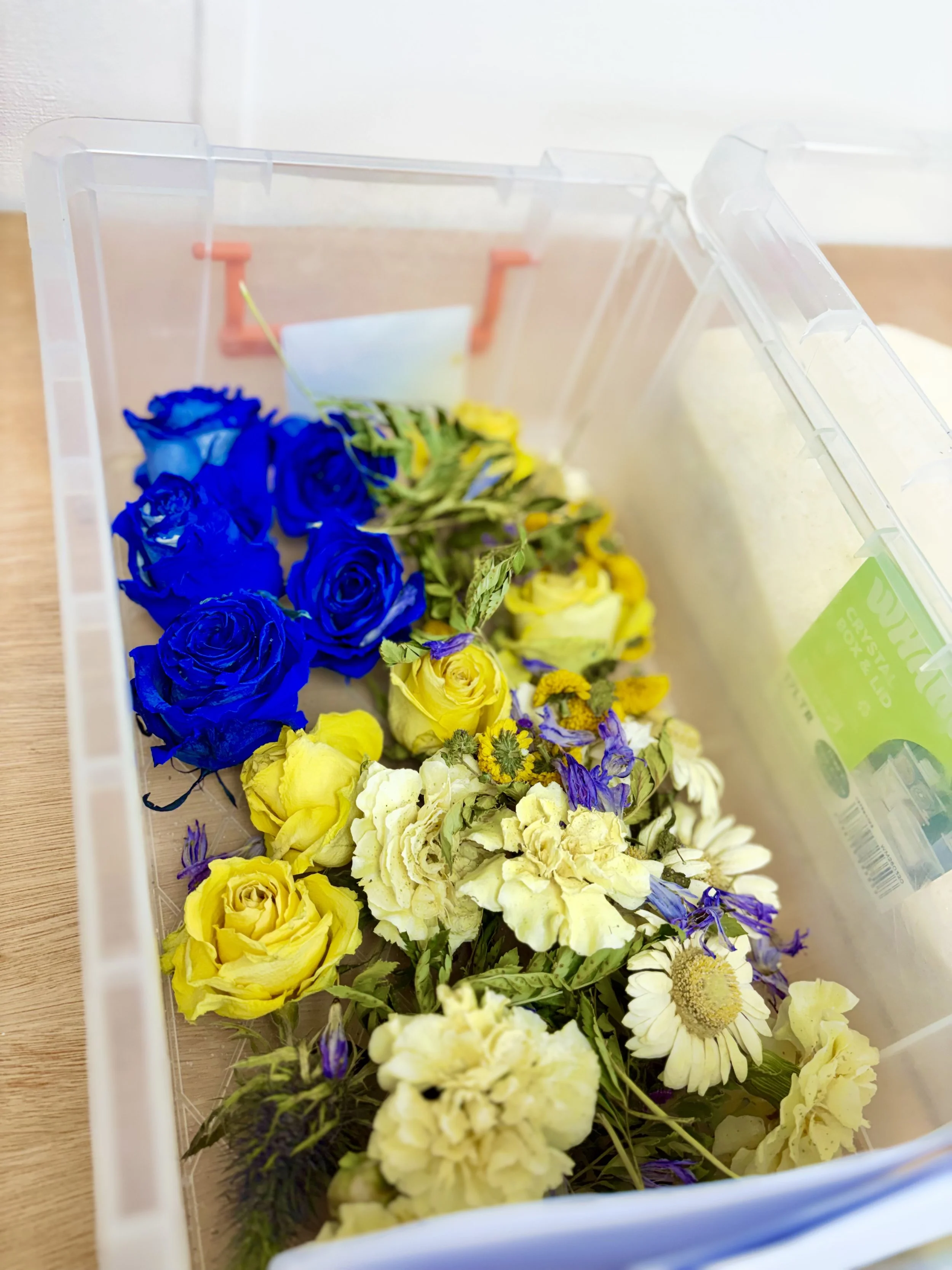A plastic container filled with a variety of fresh flowers, including yellow and blue roses, and other assorted blossoms.
