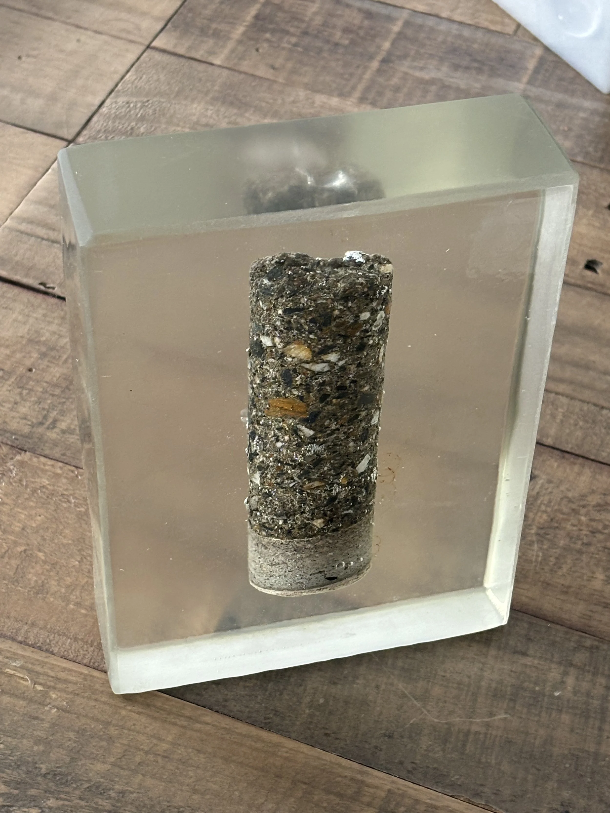 A cylindrical piece of concrete with embedded stones inside a clear glass display case, placed on a wooden surface.
