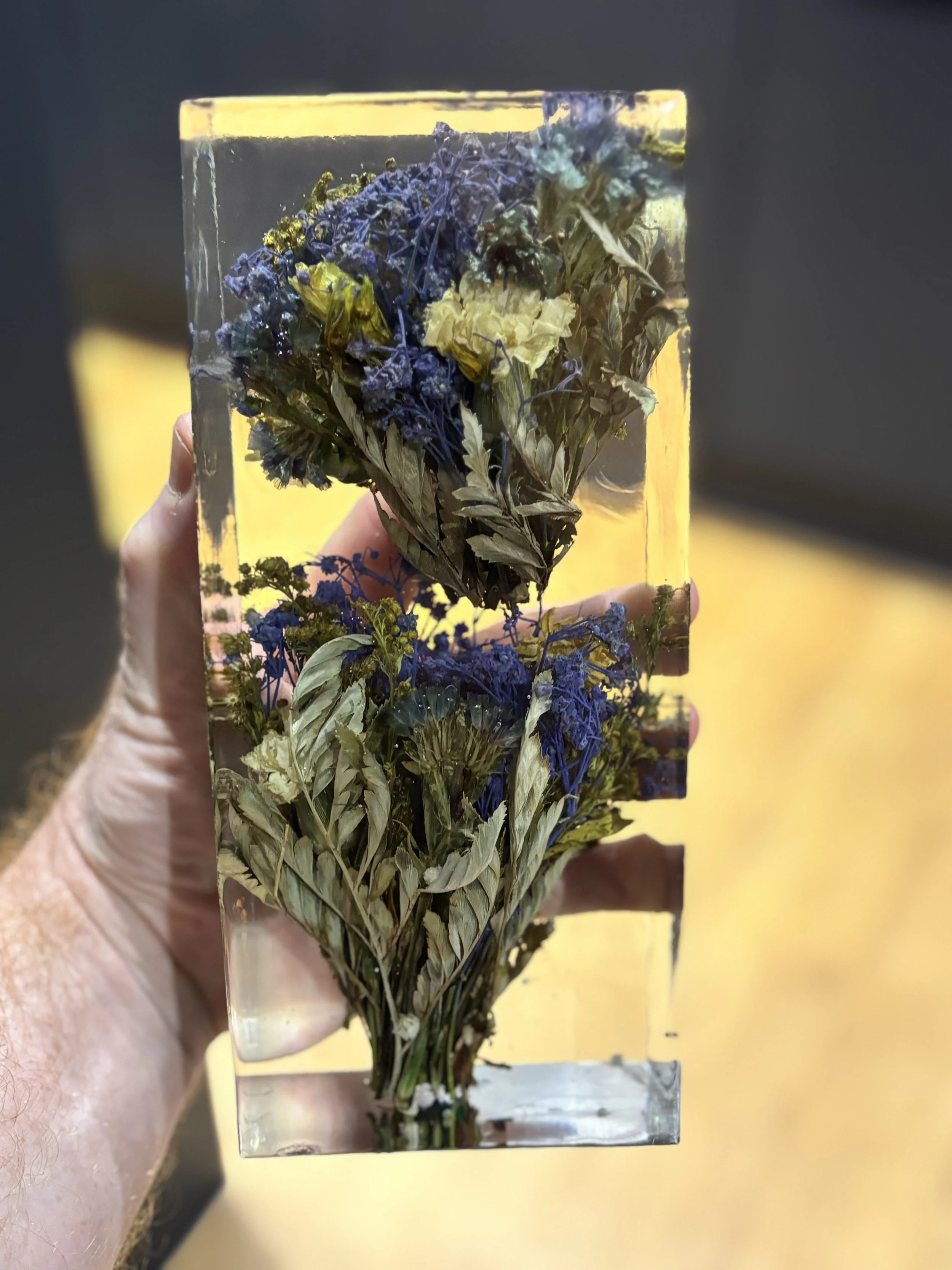 A hand holding a rectangular glass block with dried blue and yellow flowers embedded inside.