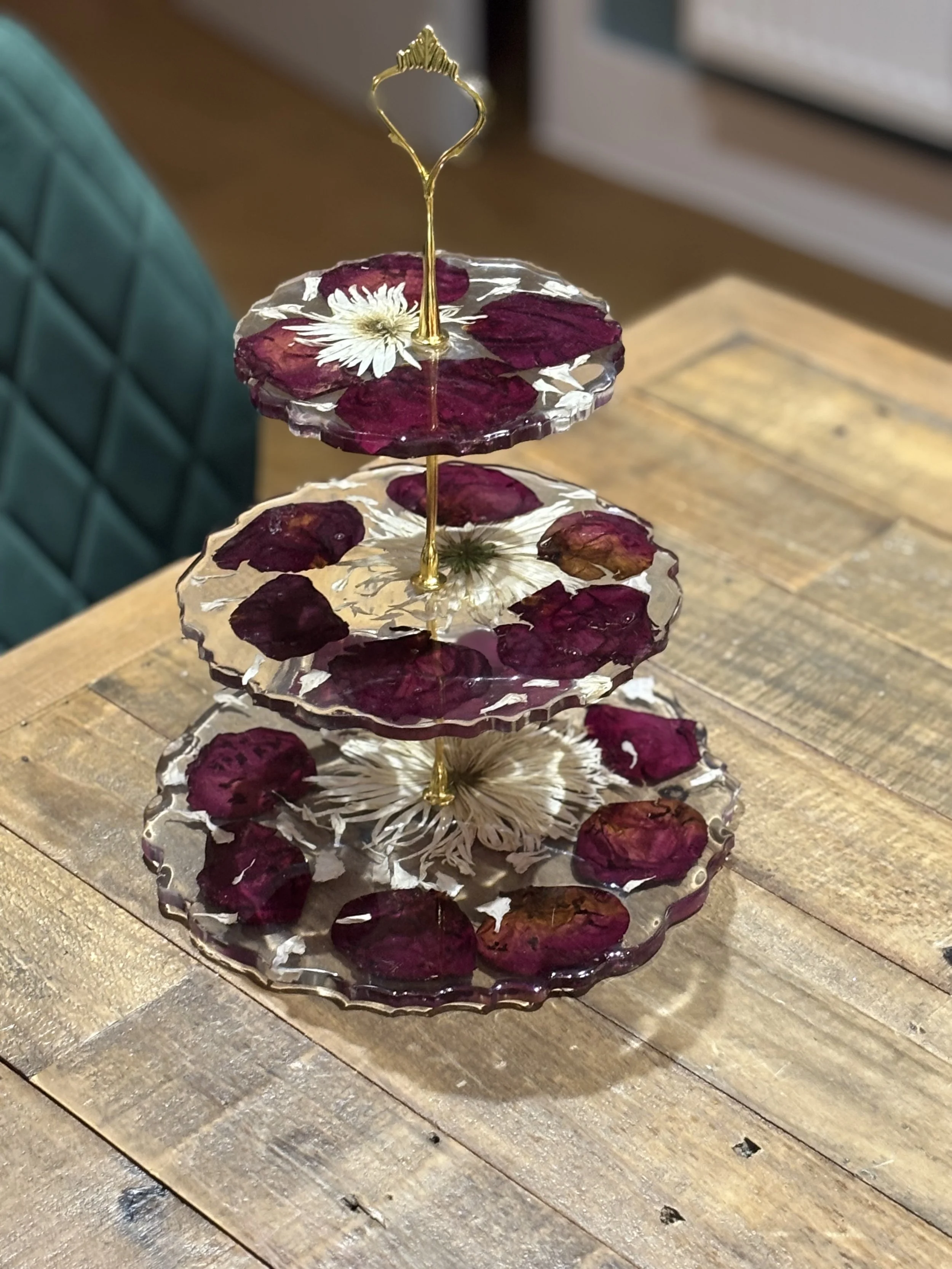 A three-tiered glass plate holder with dried rose petals and white flowers inside each tier, placed on a wooden table.