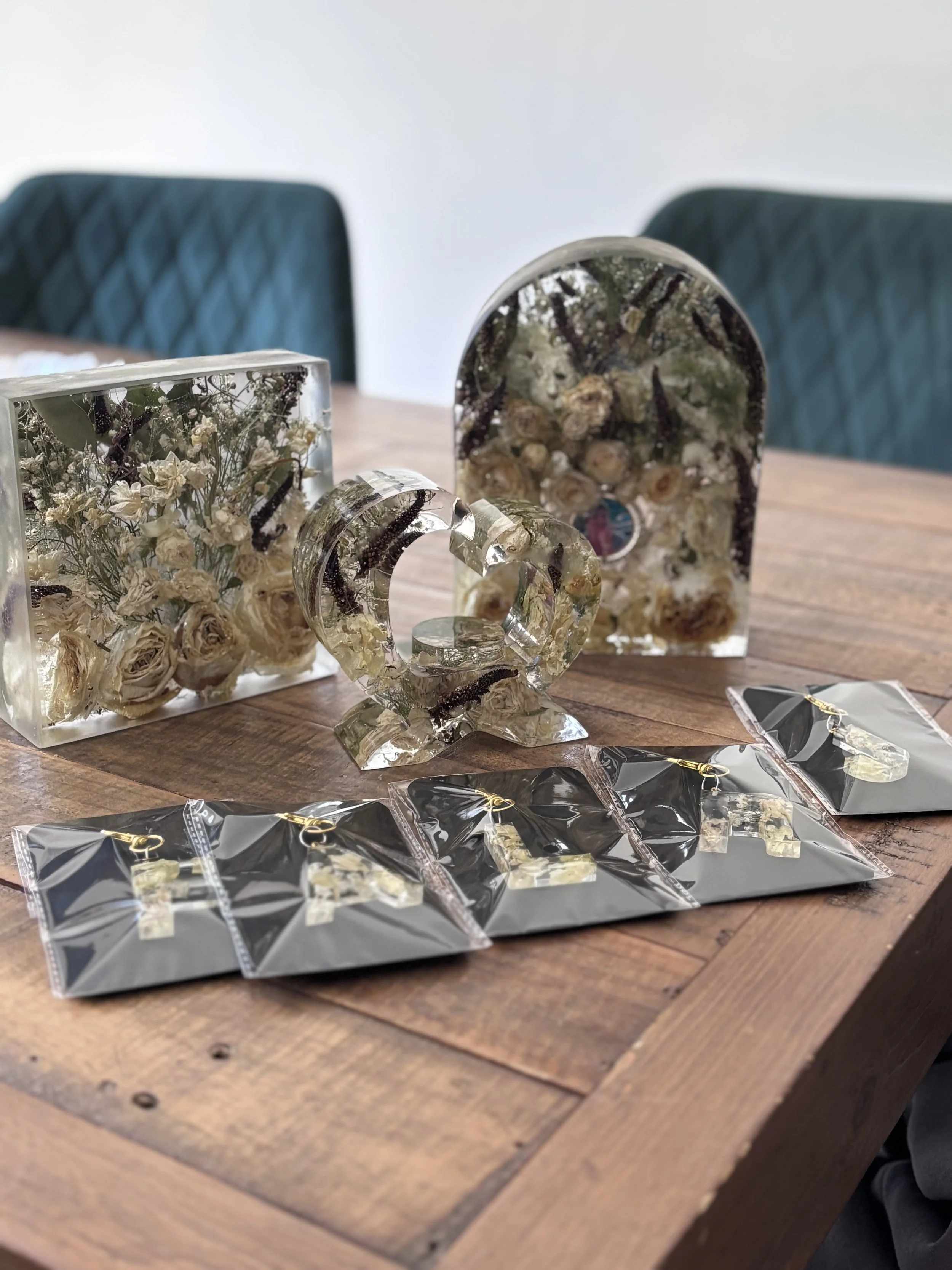 Decorative acrylic display on a wooden table, containing dried flowers and a face inside the larger piece, with gift items or jewelry in plastic packaging at the front.