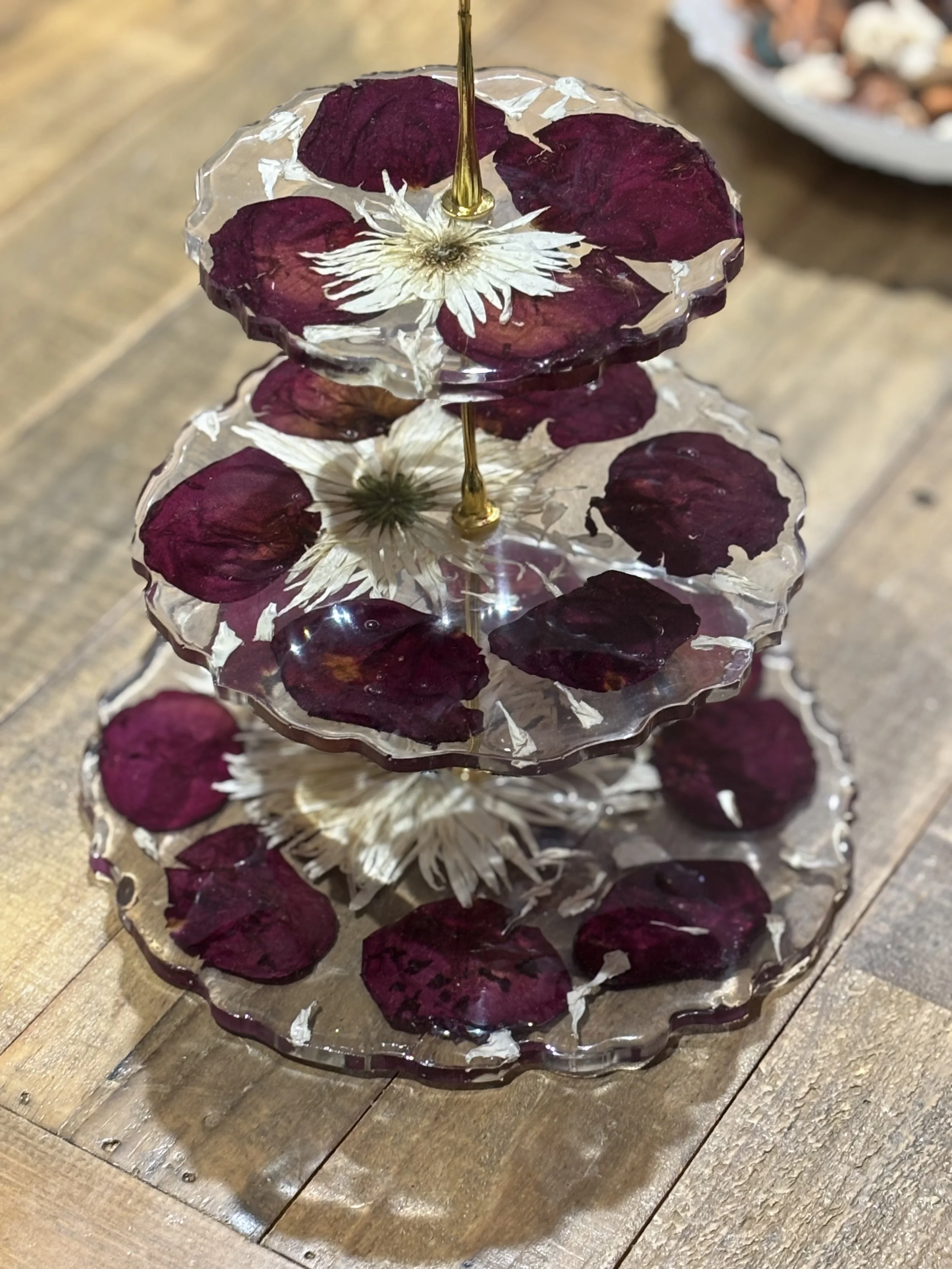 Three-tiered glass stand with dried flowers and petals inside, containing layers of burgundy petals and white flowers, placed on a wooden surface.
