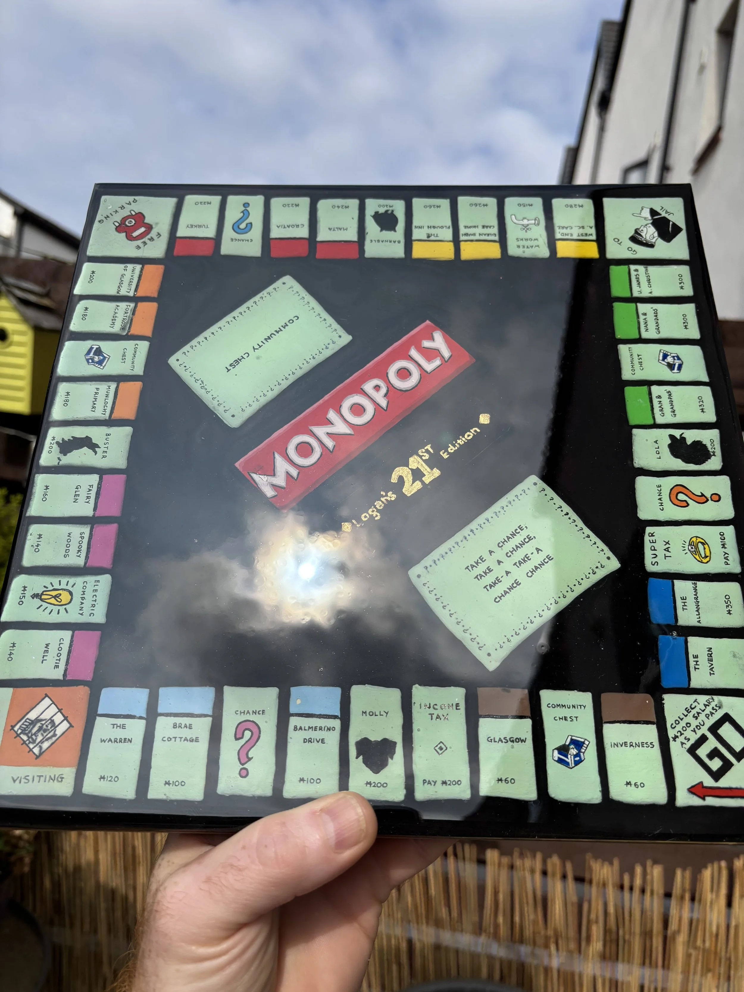 Photo of a Monopoly game board held outdoors, showing spaces such as The Warren, Brae Cottage, Chance, Balmerino Drive, Incense Tax, Glasgow, Community Chest, Inverness, and GO to Collect $200.