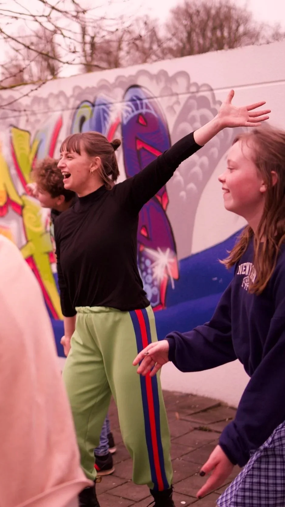 Junge Frauen lachen und tanzen vor einer mit Graffiti bemalten Wand bei Nacht.