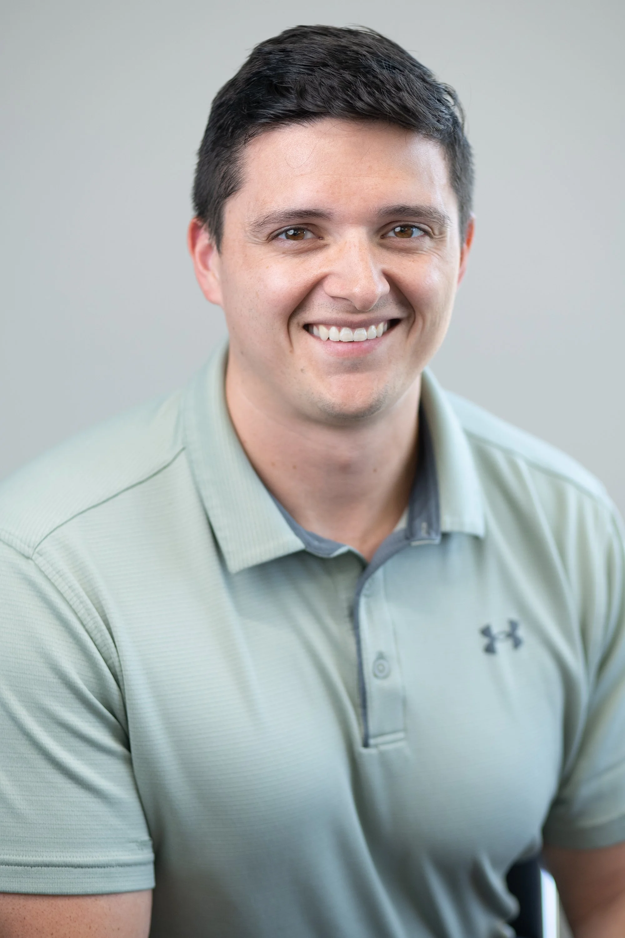 Close-up portrait of a smiling young man with short dark hair, wearing a light green Under Armour polo shirt, against a plain grey background.
