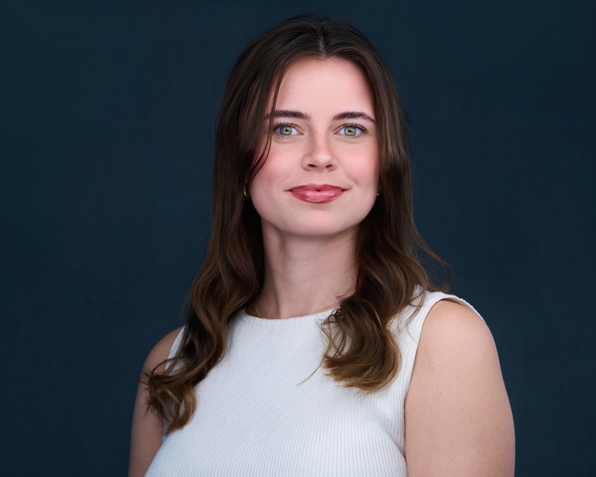 A woman with long brown hair, blue eyes, and fair skin, wearing a sleeveless white top, smiling against a dark blue background.
