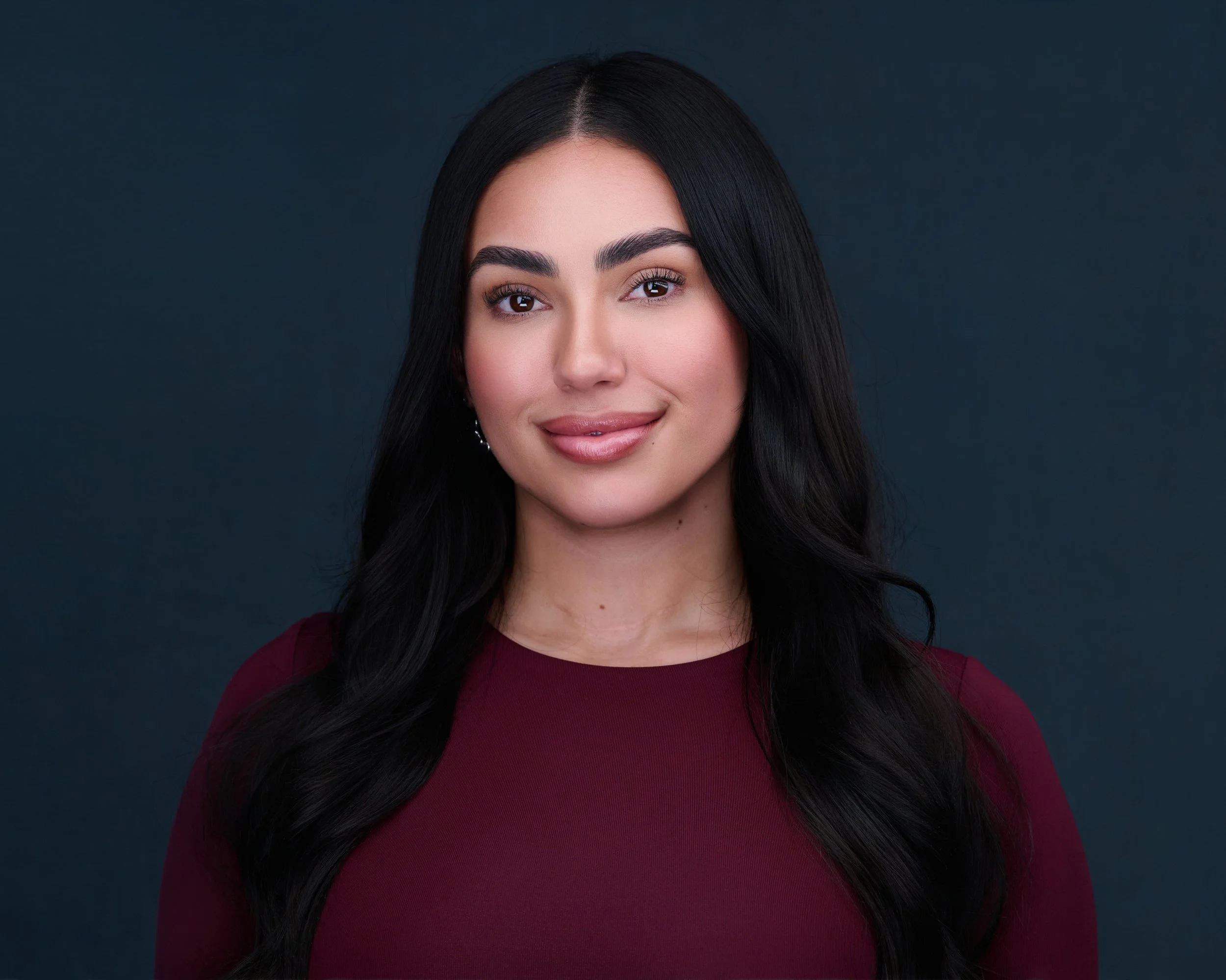 A woman with long black hair, wearing a maroon top, smiling slightly, against a dark background.