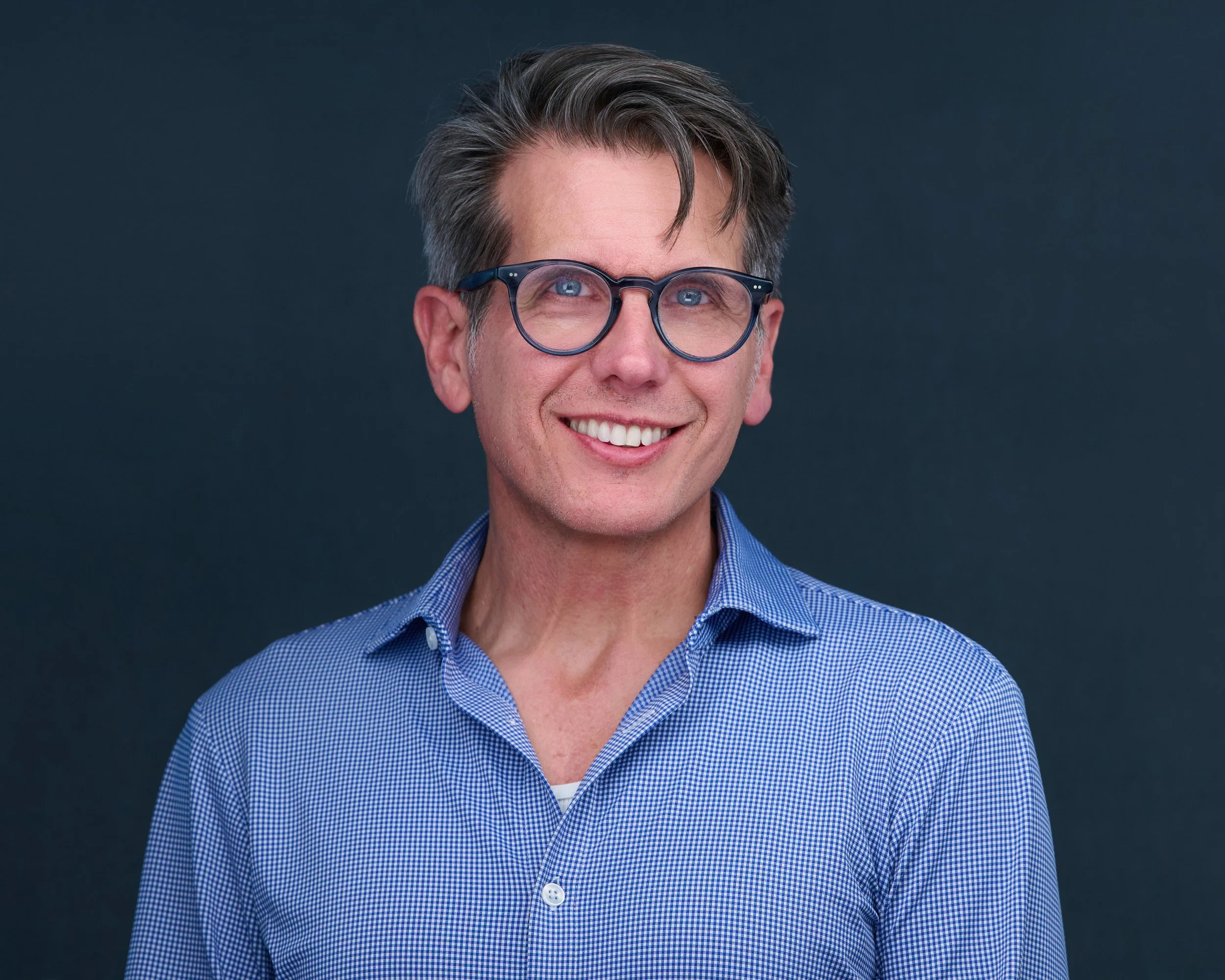Smiling man with glasses wearing a blue checkered shirt against a dark background.