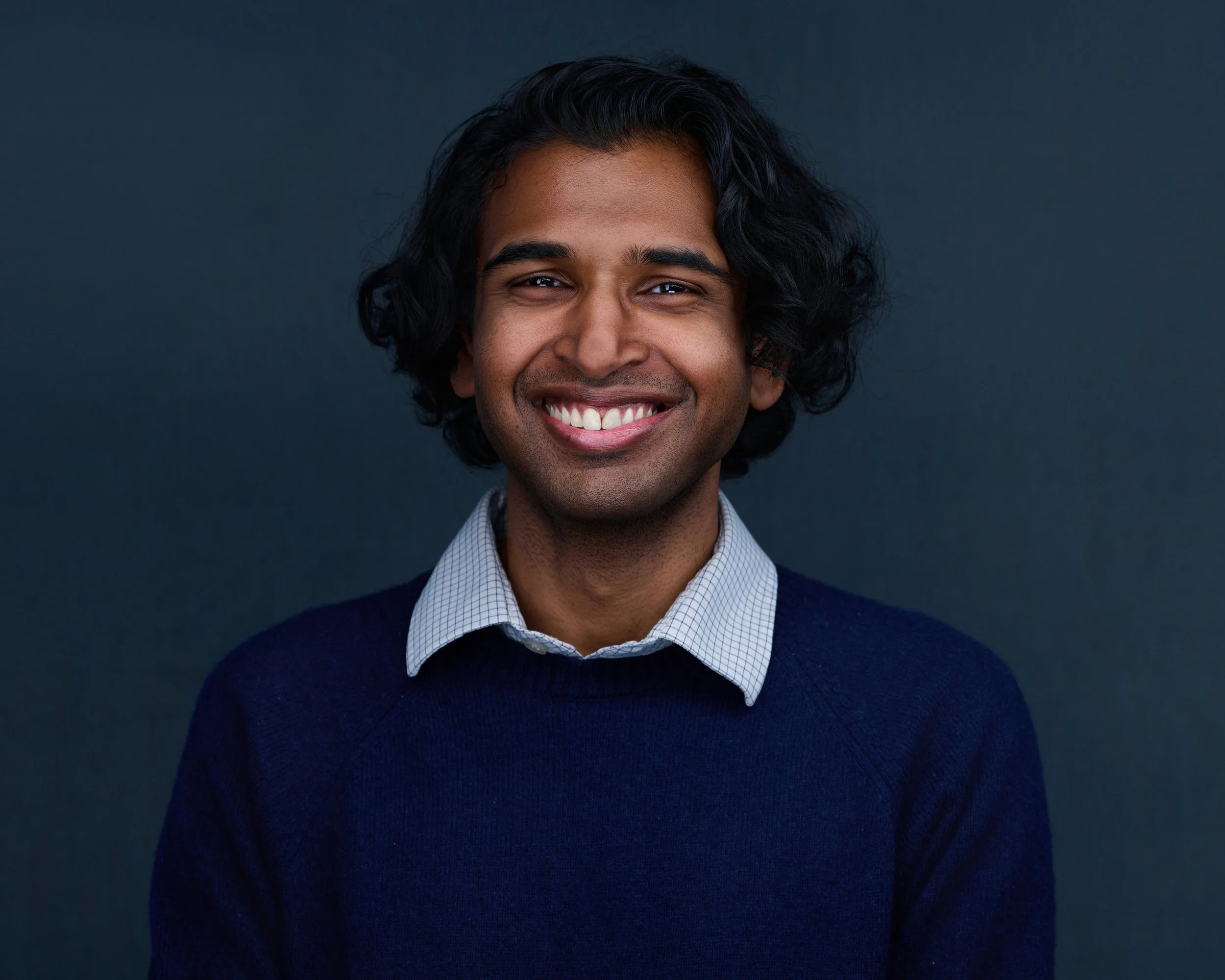 A man with dark curly hair and medium brown skin, smiling, wearing a blue sweater over a white collared shirt, against a dark background.