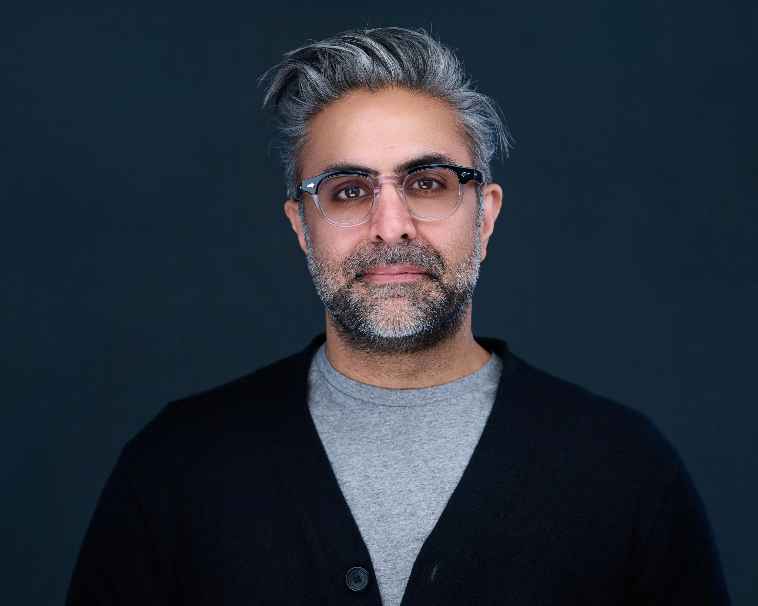 Portrait of a middle-aged man with gray hair, beard, wearing glasses, a gray shirt, and a black cardigan against a dark background.