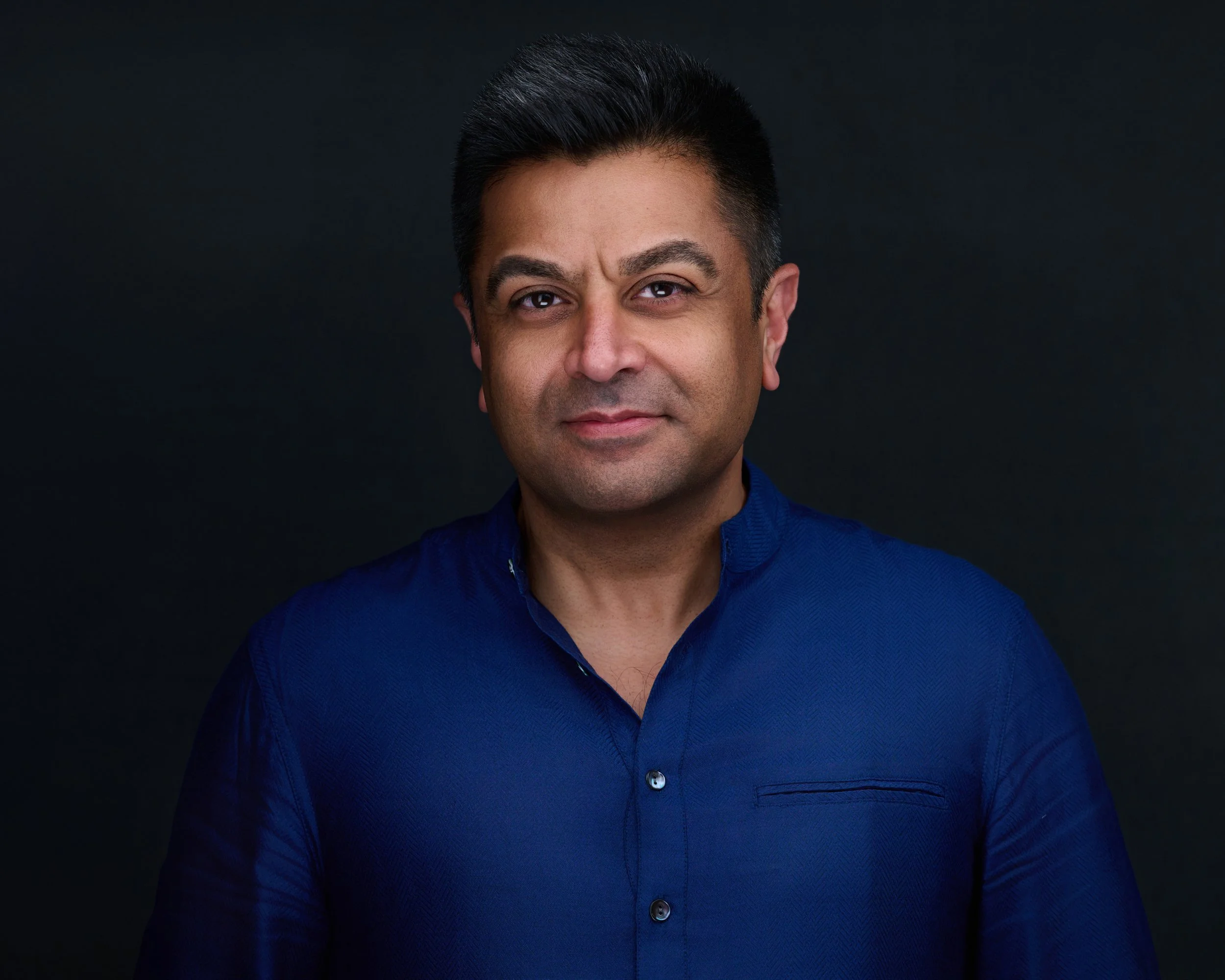A man with short black hair, wearing a dark blue shirt, looking at the camera with a slight smile, against a dark background.