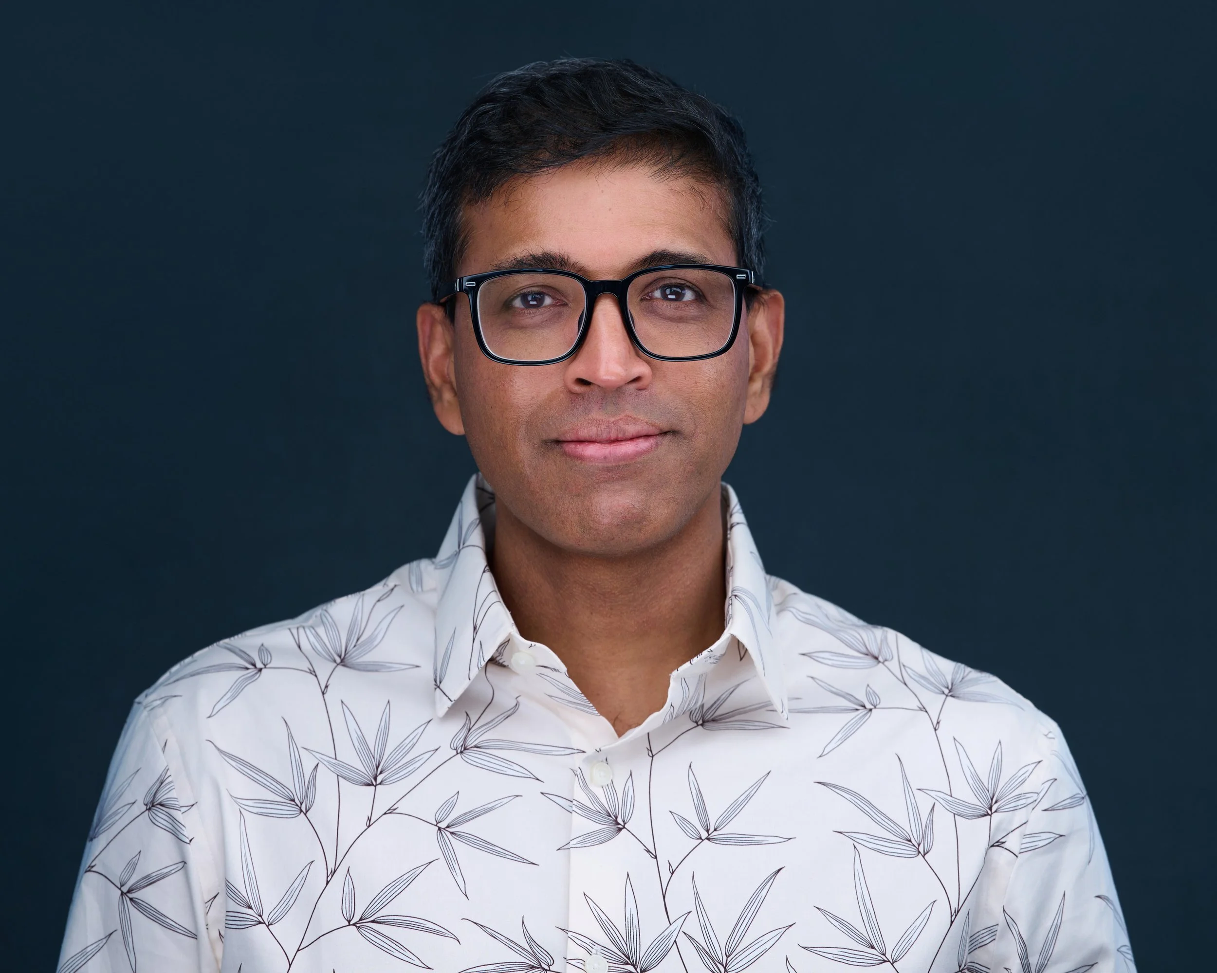 Headshot of a man with short dark hair and glasses, wearing a white shirt with a leaf pattern, standing against a dark background.