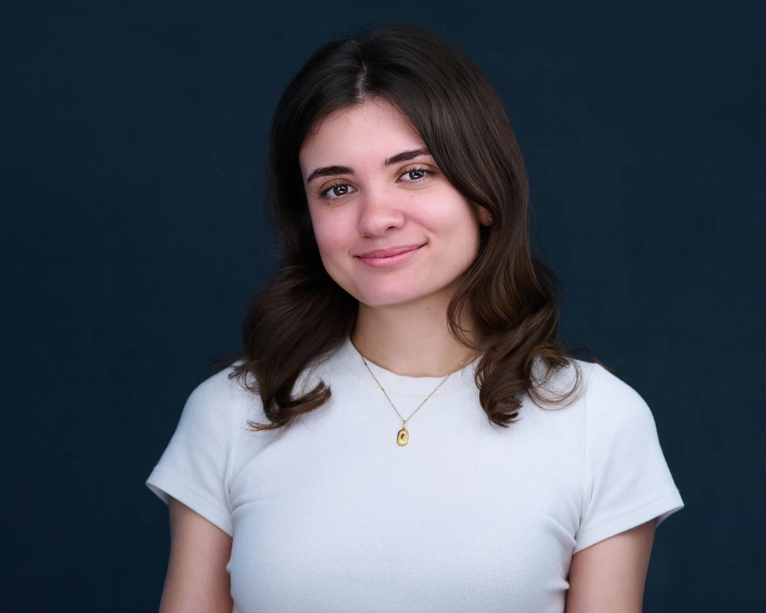 A young woman with dark hair wearing a white t-shirt and a gold necklace, smiling softly against a dark background.