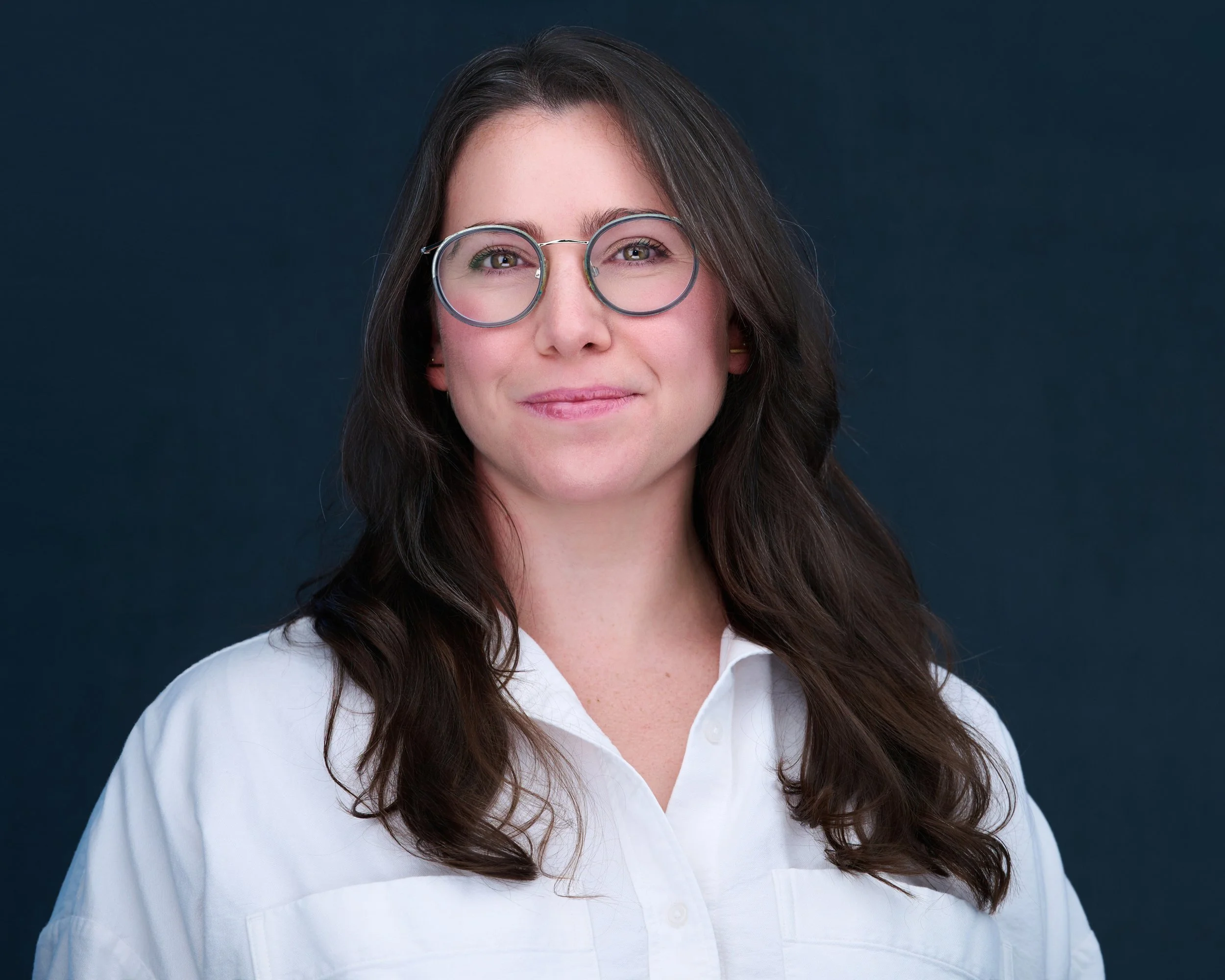 A woman with long dark hair, wearing glasses and a white shirt, smiling against a dark background.
