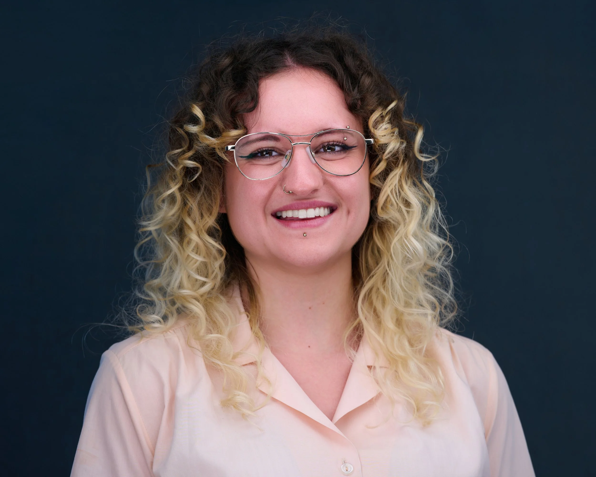 A woman with curly blonde hair, wearing glasses and a light pink blouse, smiling in front of a dark background.
