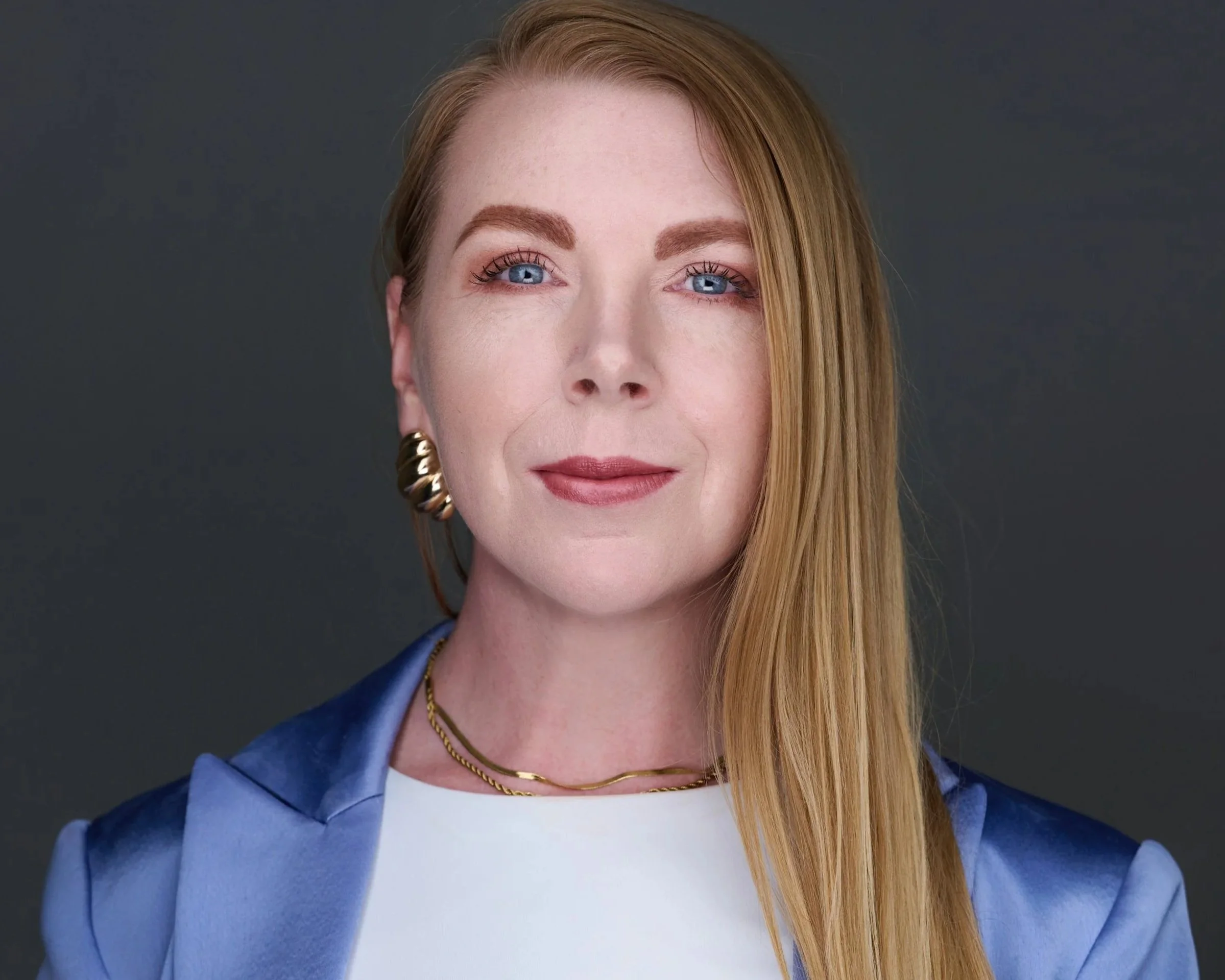 Close-up of a woman with long red hair, blue eyes, wearing a blue blazer, white top, gold jewelry, and light makeup, smiling softly against a dark background.
