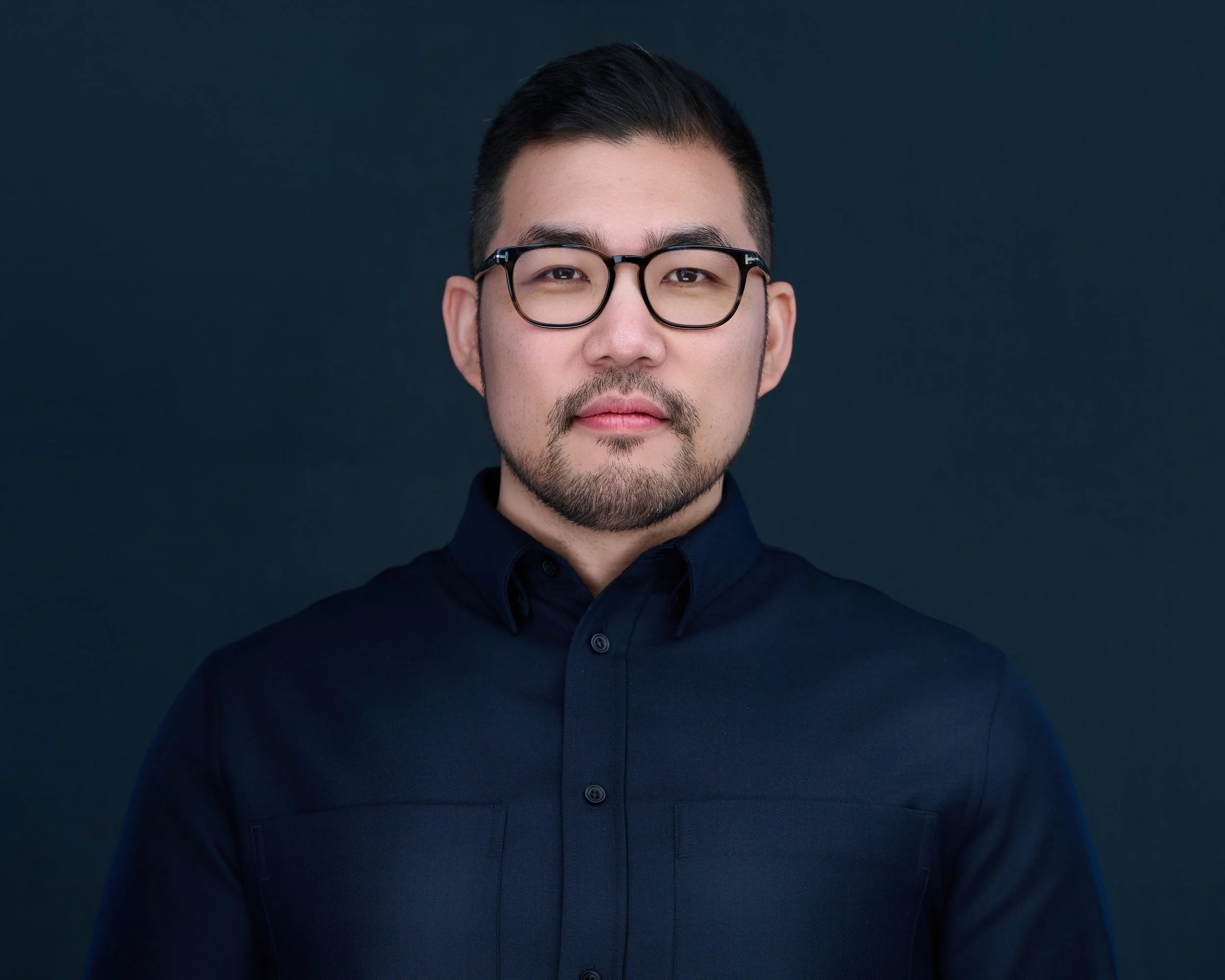 Portrait of a man with glasses and a dark collared shirt against a dark background.