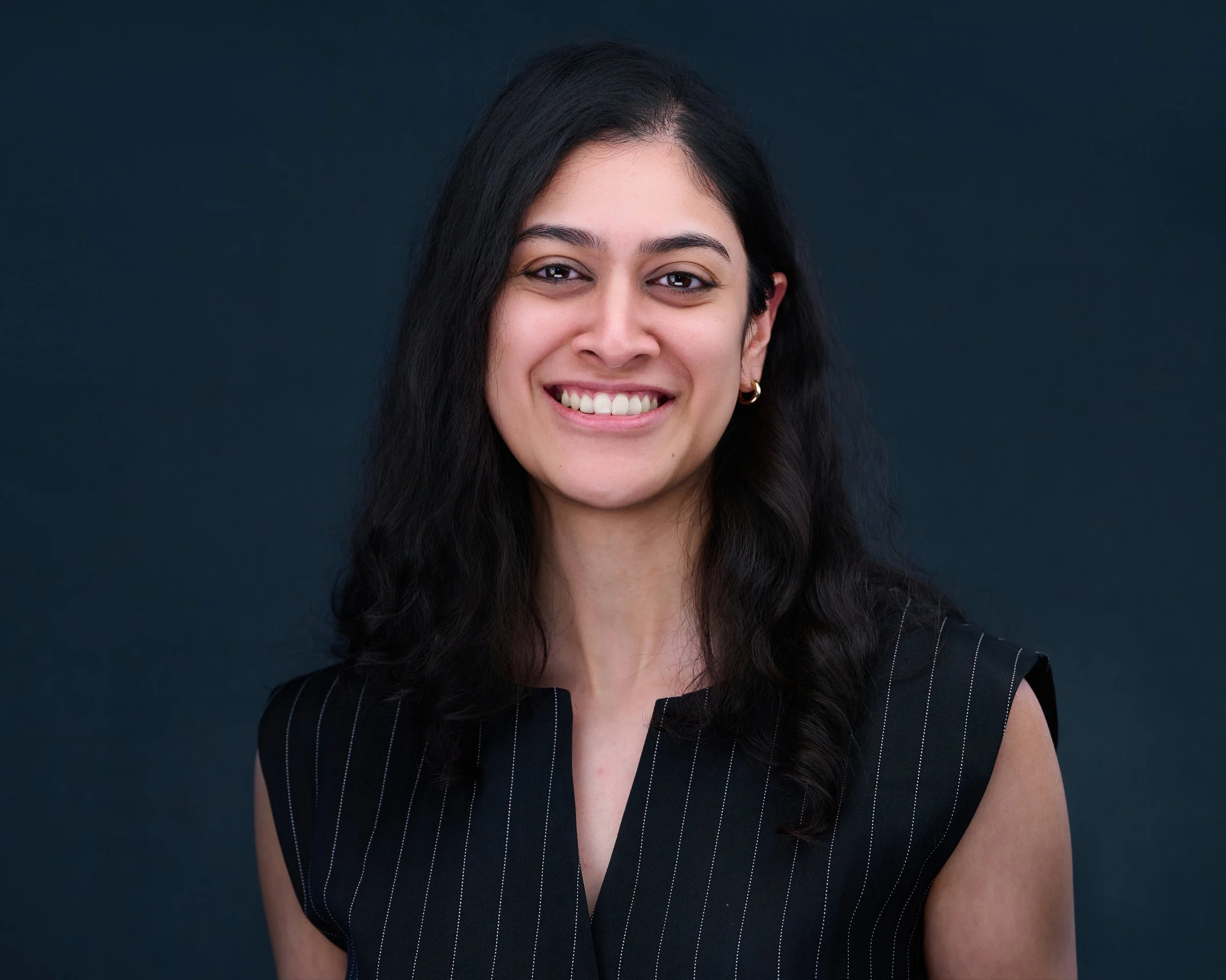 A young woman with dark wavy hair, wearing a black sleeveless top with white pinstripes, smiling against a dark blue background.