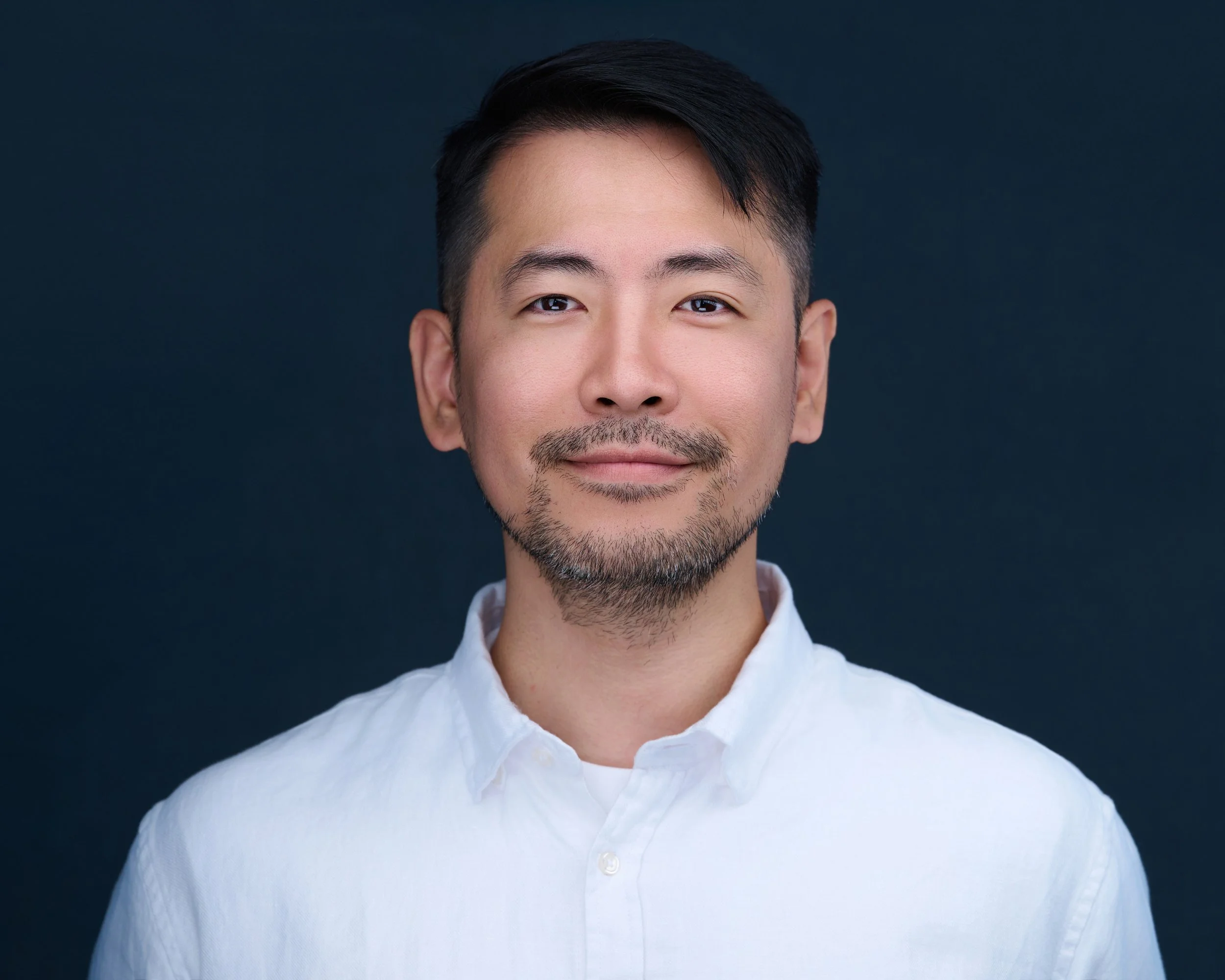 Portrait of a man with short black hair, facial hair, wearing a white button-up shirt, smiling subtly against a dark background.
