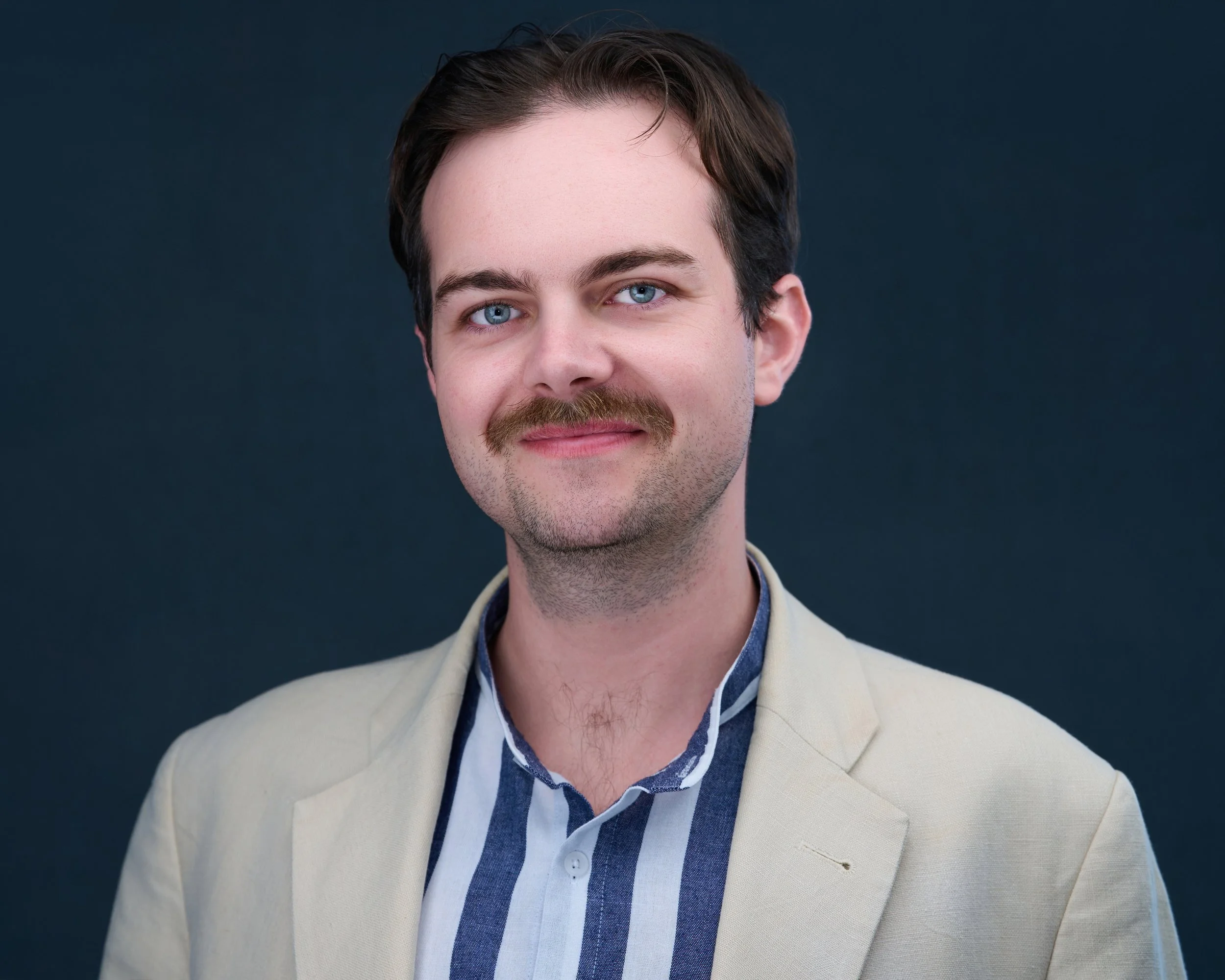 A young man with blue eyes, brown hair, and a mustache wearing a beige blazer and a striped shirt, looking at the camera with a slight smile against a dark background.