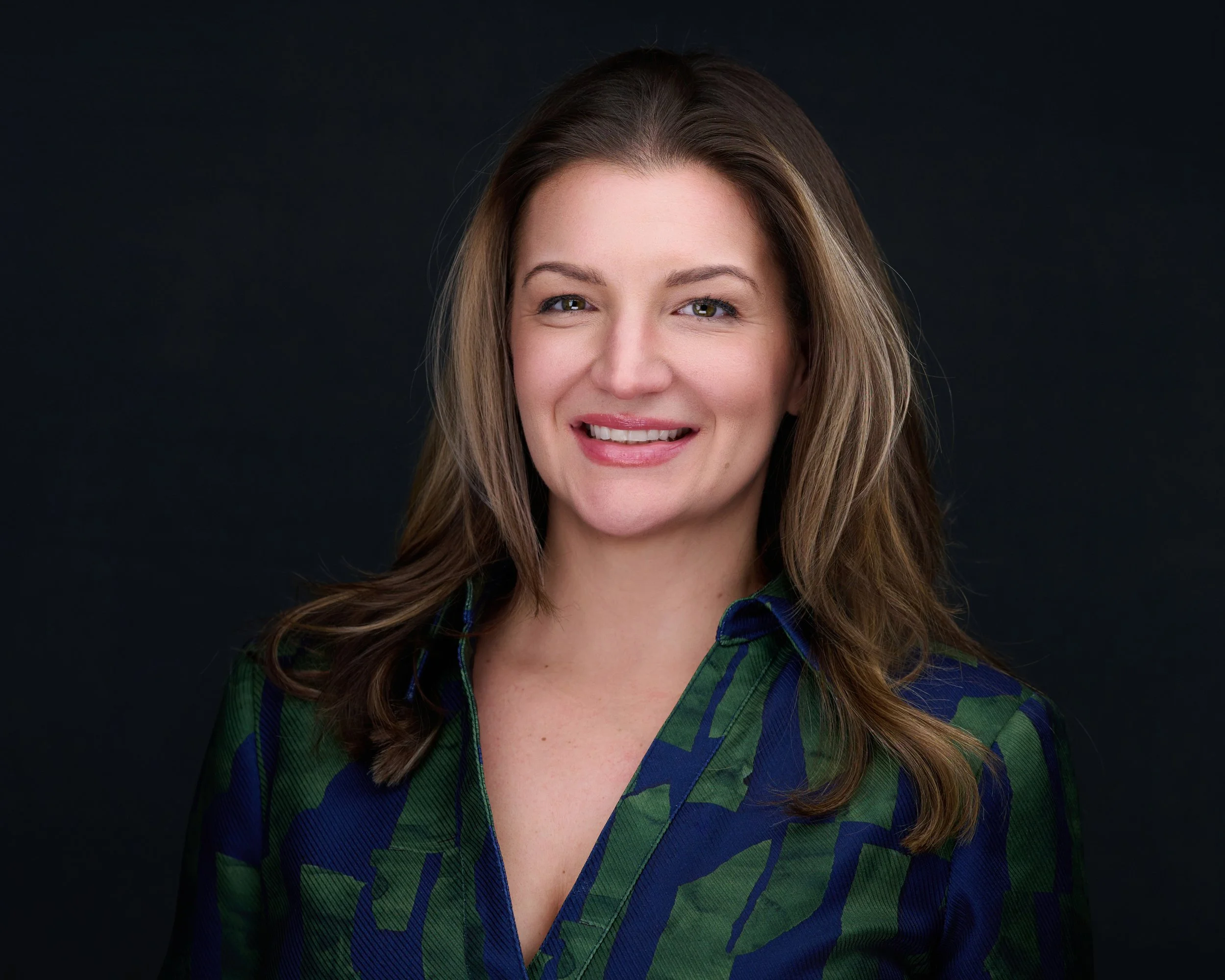 Portrait of a woman with shoulder-length brown hair, smiling, wearing a multicolored patterned blouse, against a dark background.