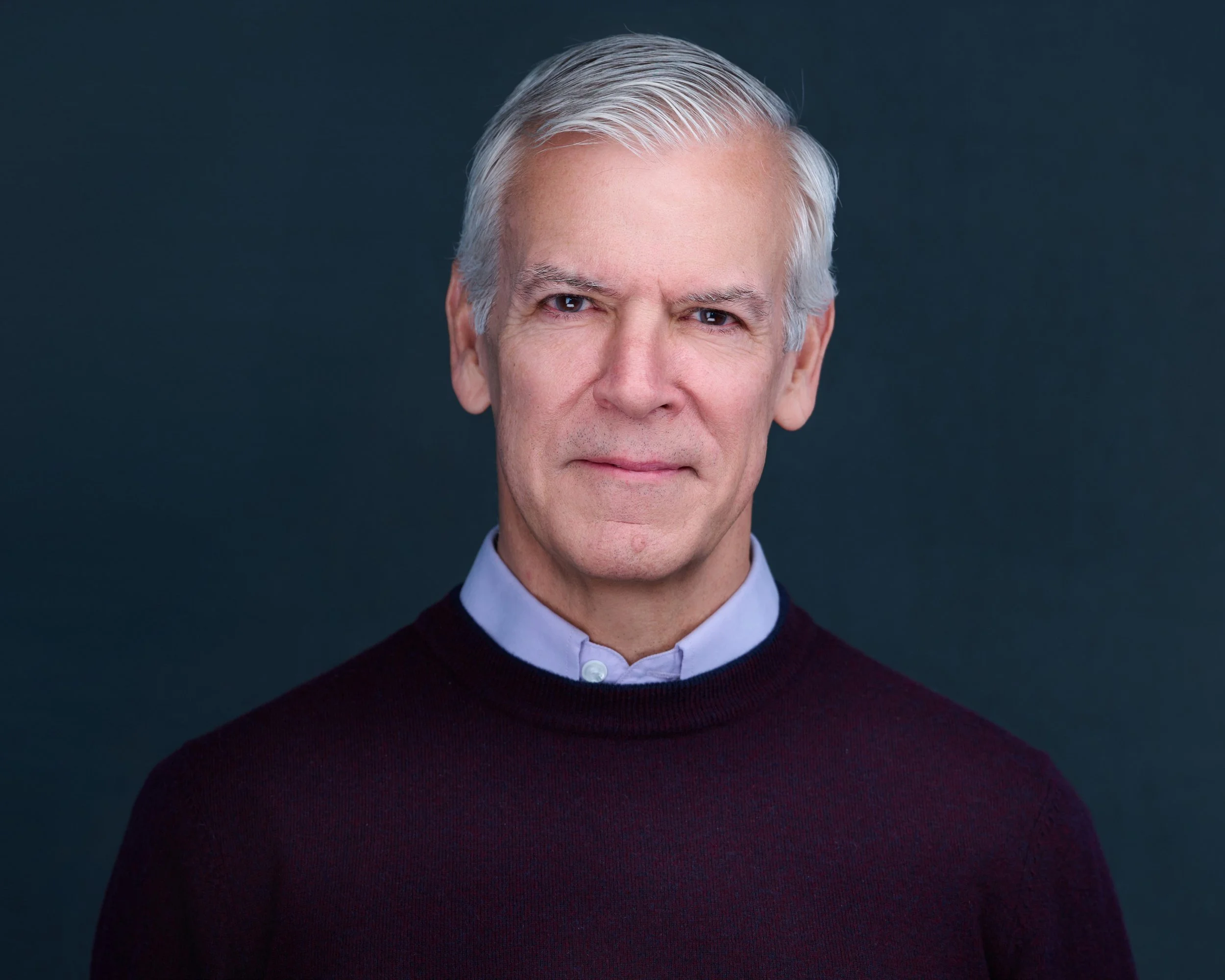 Portrait of an older man with white hair, wearing a light blue collared shirt under a dark sweater, standing against a dark background.