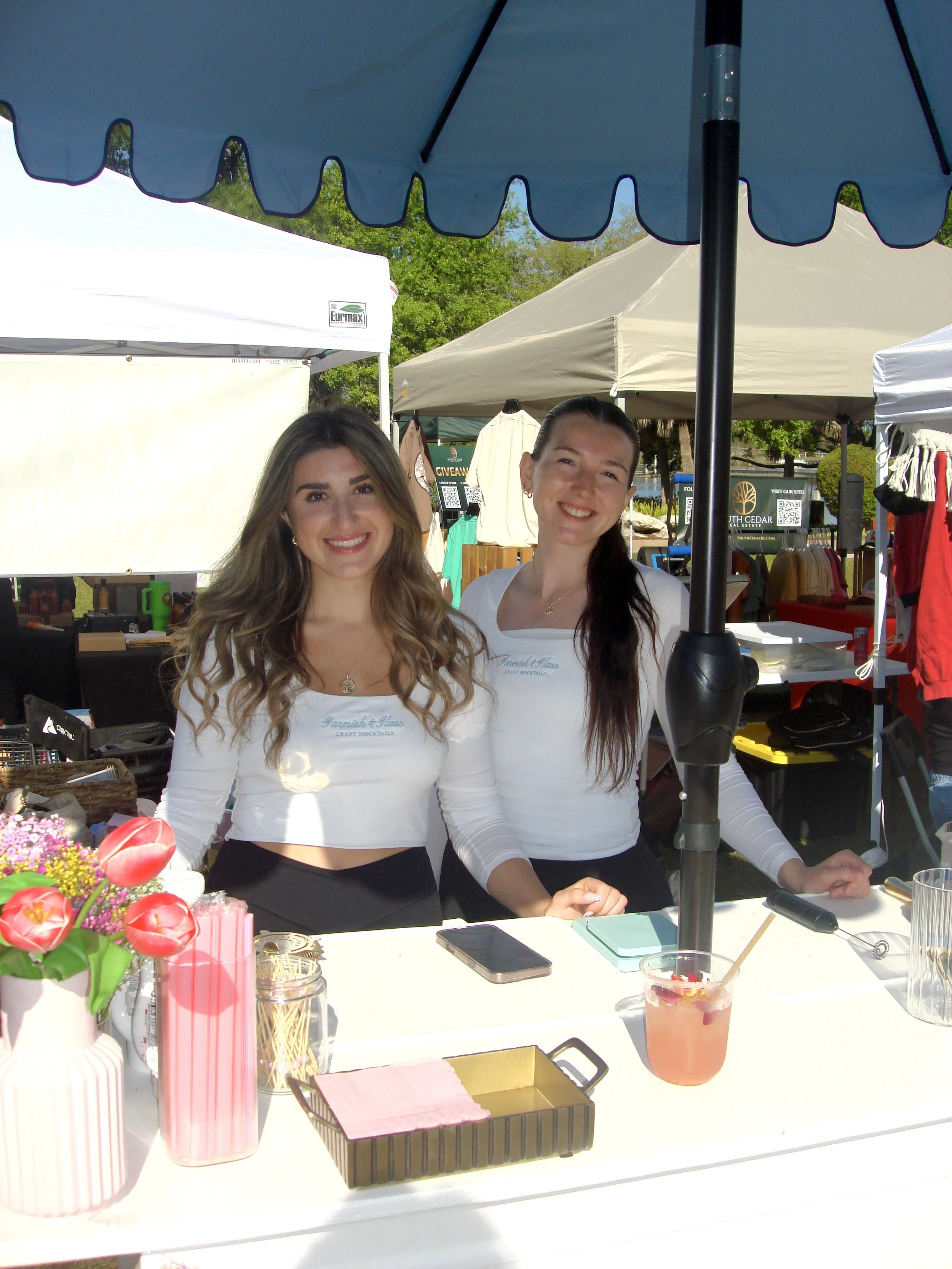 Two women behind a vendor table at an outdoor market. They are smiling, wearing white long-sleeve shirts with a logo, and standing under a blue canopy. The table has flowers, a pitcher, and a drink, with other tents and market stalls visible in the background.