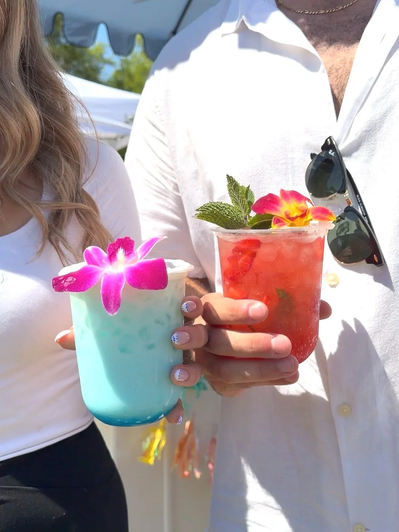 Close-up of two people holding colorful cocktails with garnishes, ice, and flowers, outdoors at a sunny event.