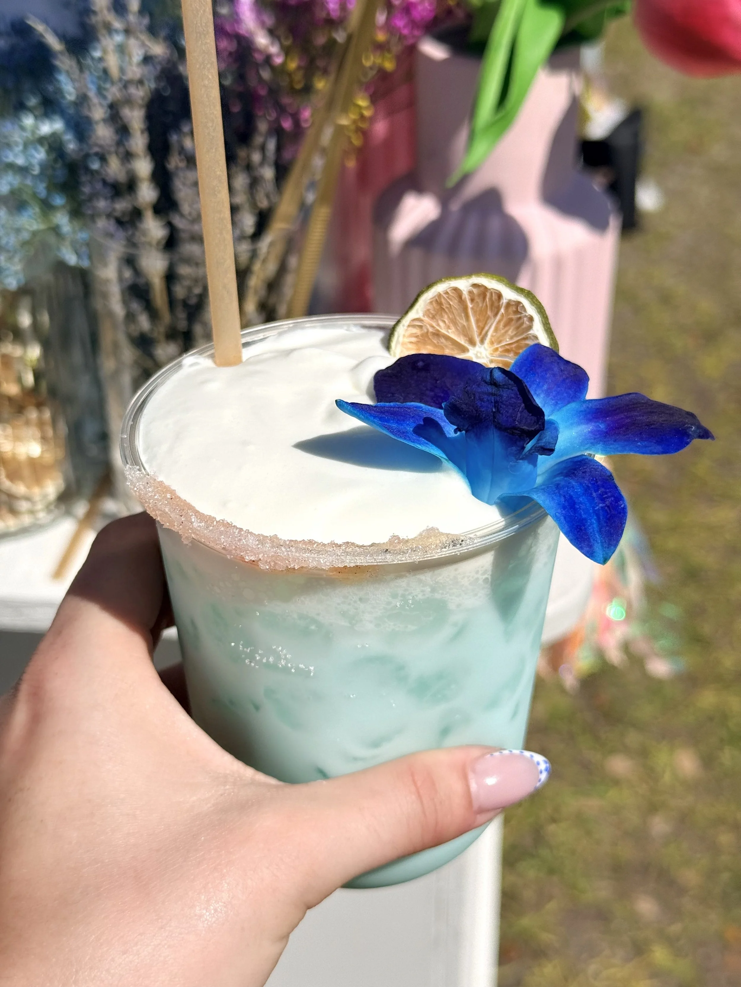 A hand holding a pastel blue cocktail garnished with a dried lemon slice and a blue flower, with a pink flower pot and green plants in the background.
