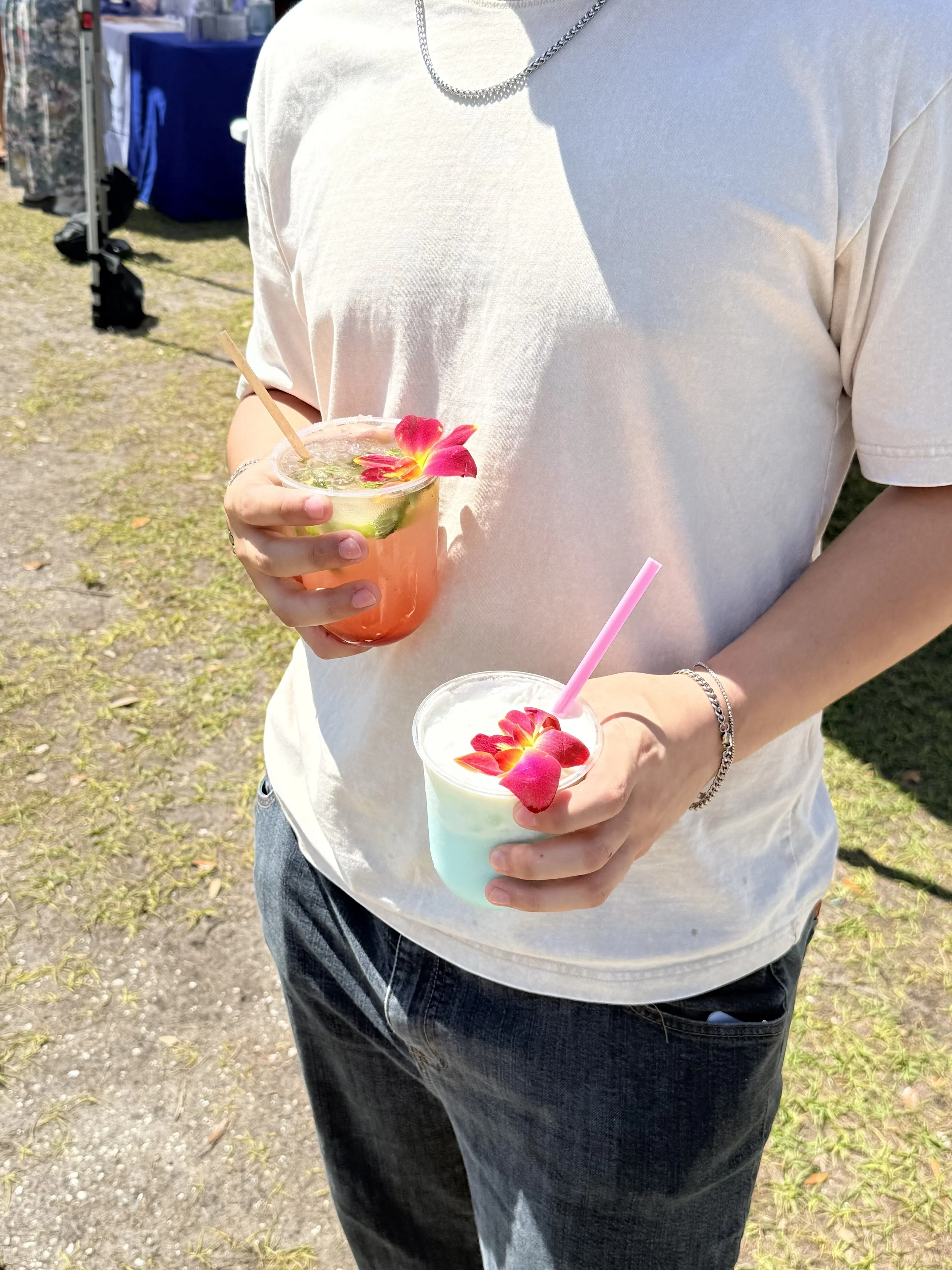 Person holding two colorful floral garnished beverages outdoors in sunny weather.