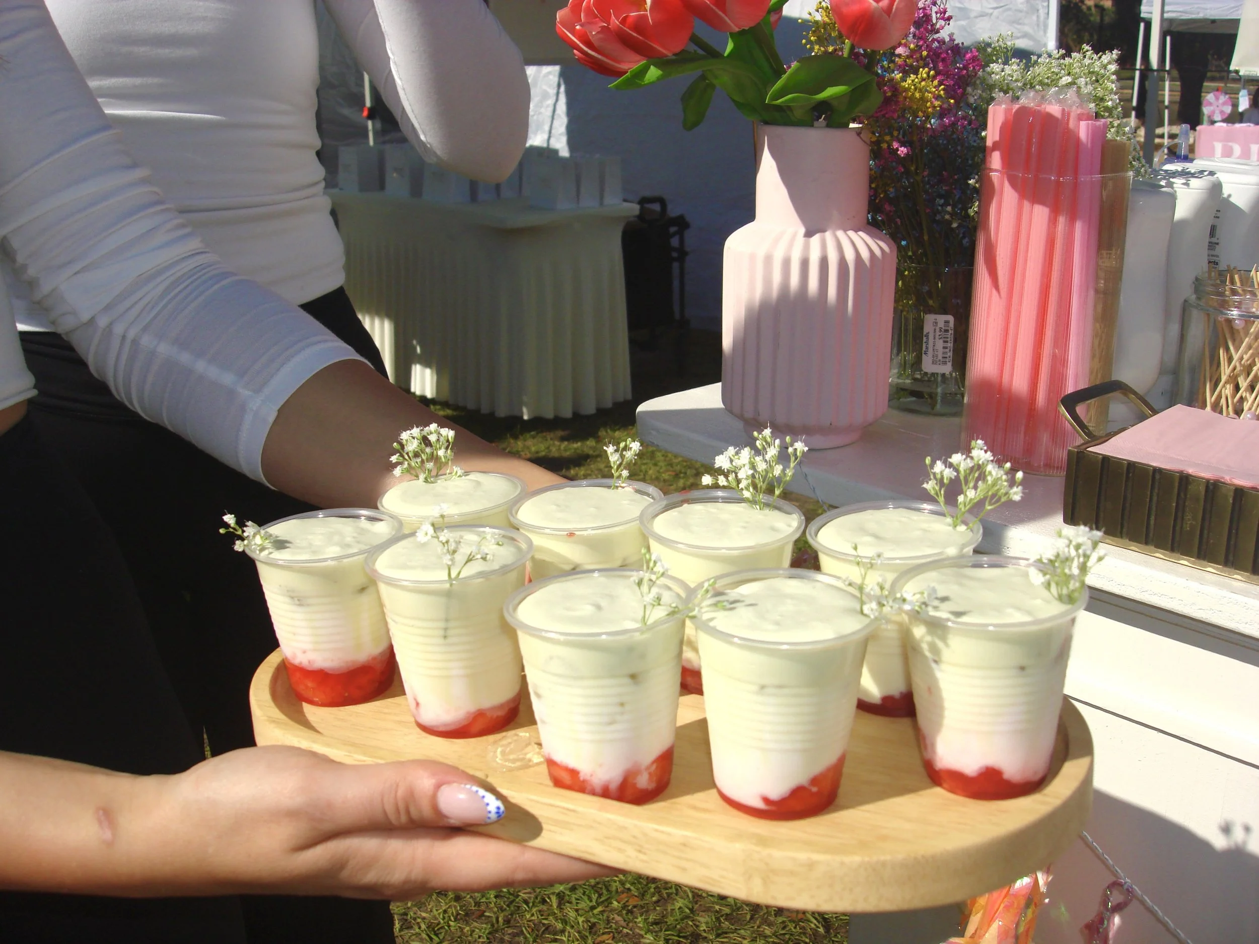 A person holding a wooden tray with small cups of layered dessert topped with small white flowers, at an outdoor event with pink vases and flowers on a table in the background.