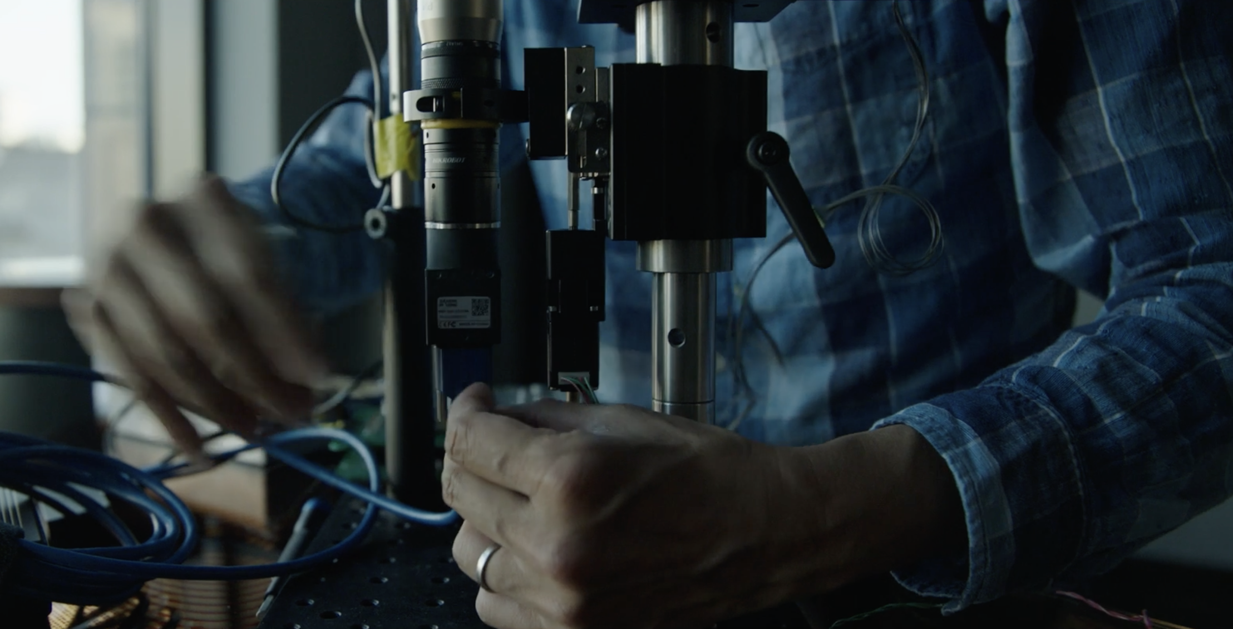 Person adjusting scientific equipment with cables and a blue plaid shirt in a laboratory setting.