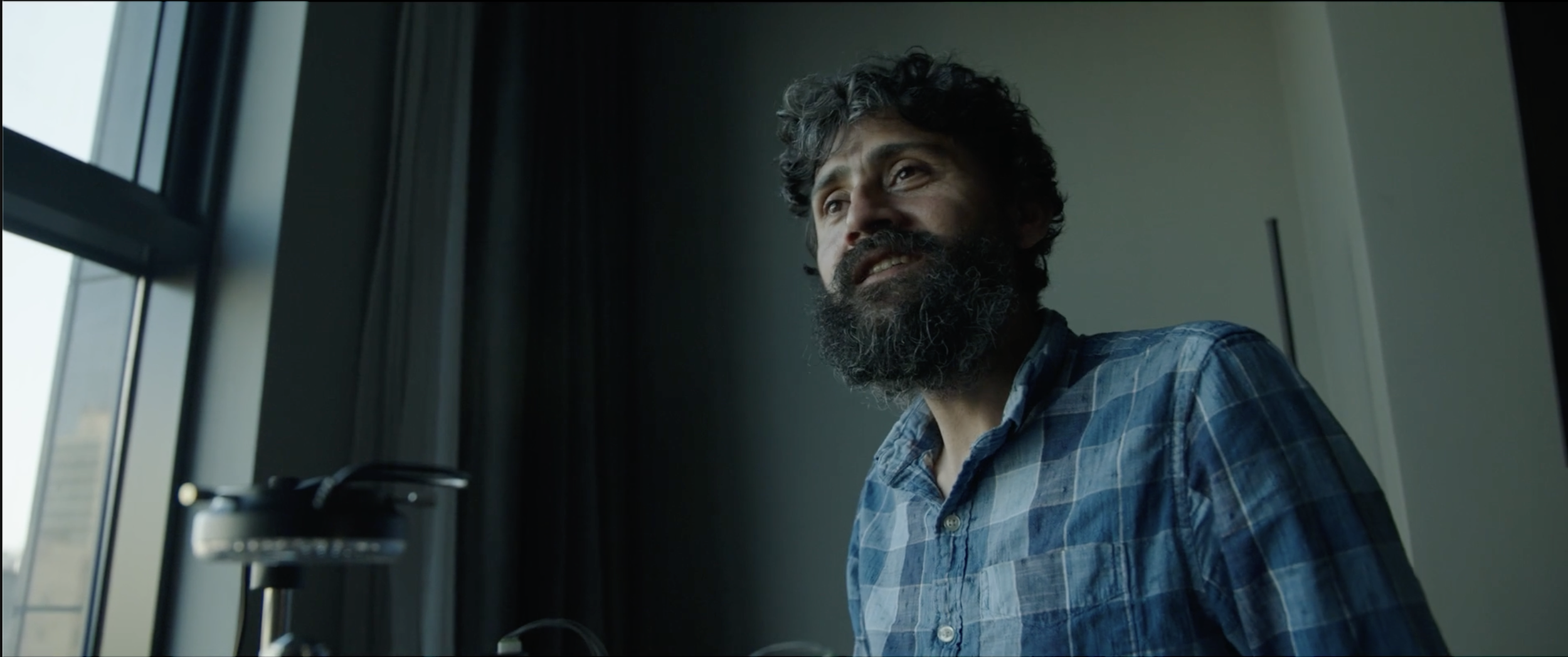 A man with a beard and curly hair wearing a blue checkered shirt, looking to the side with a thoughtful expression, standing by a window with a cityscape in the background.