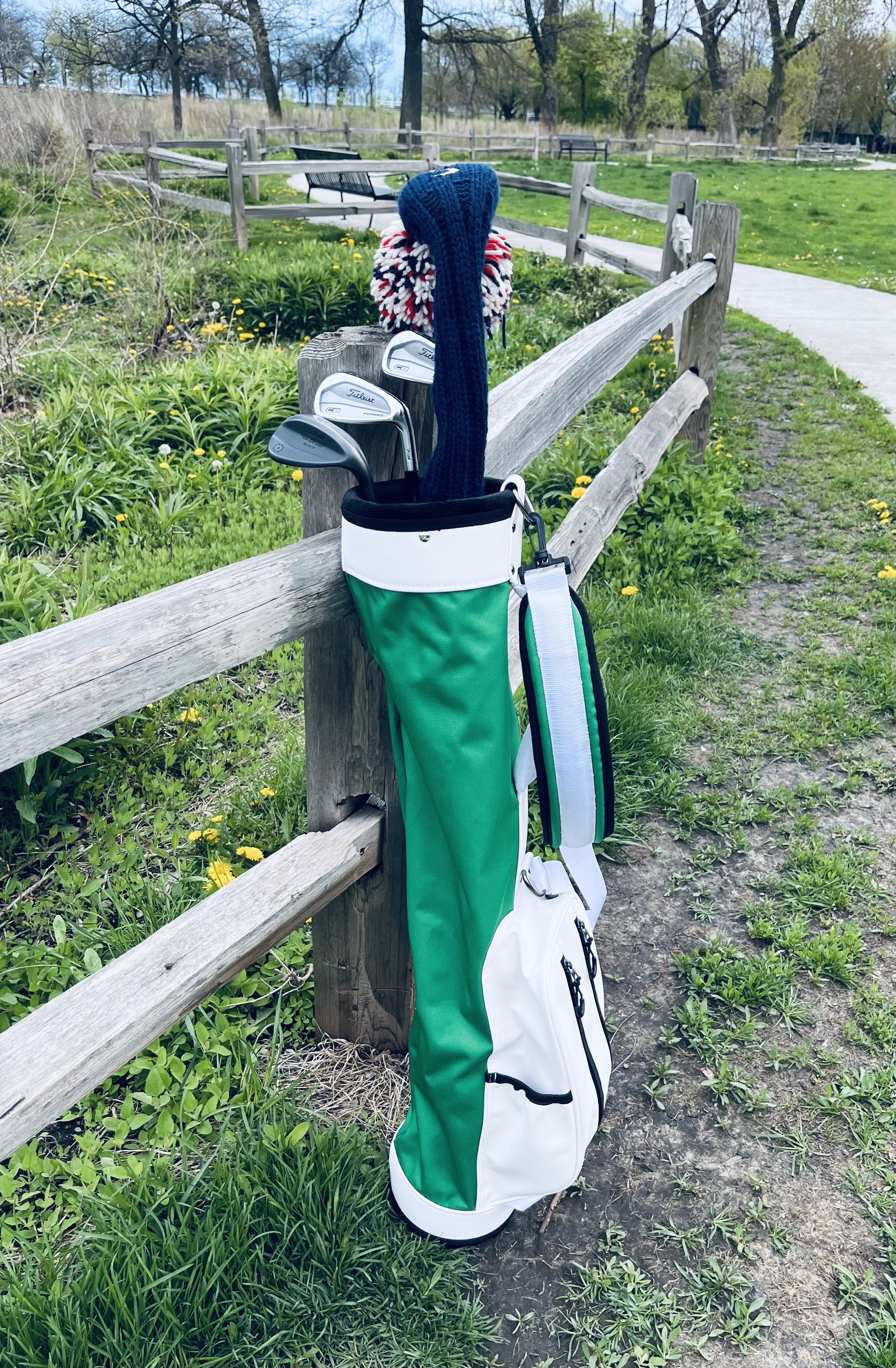 A golf bag with clubs leaning against a wooden fence in a park, with trees and a sidewalk visible in the background.