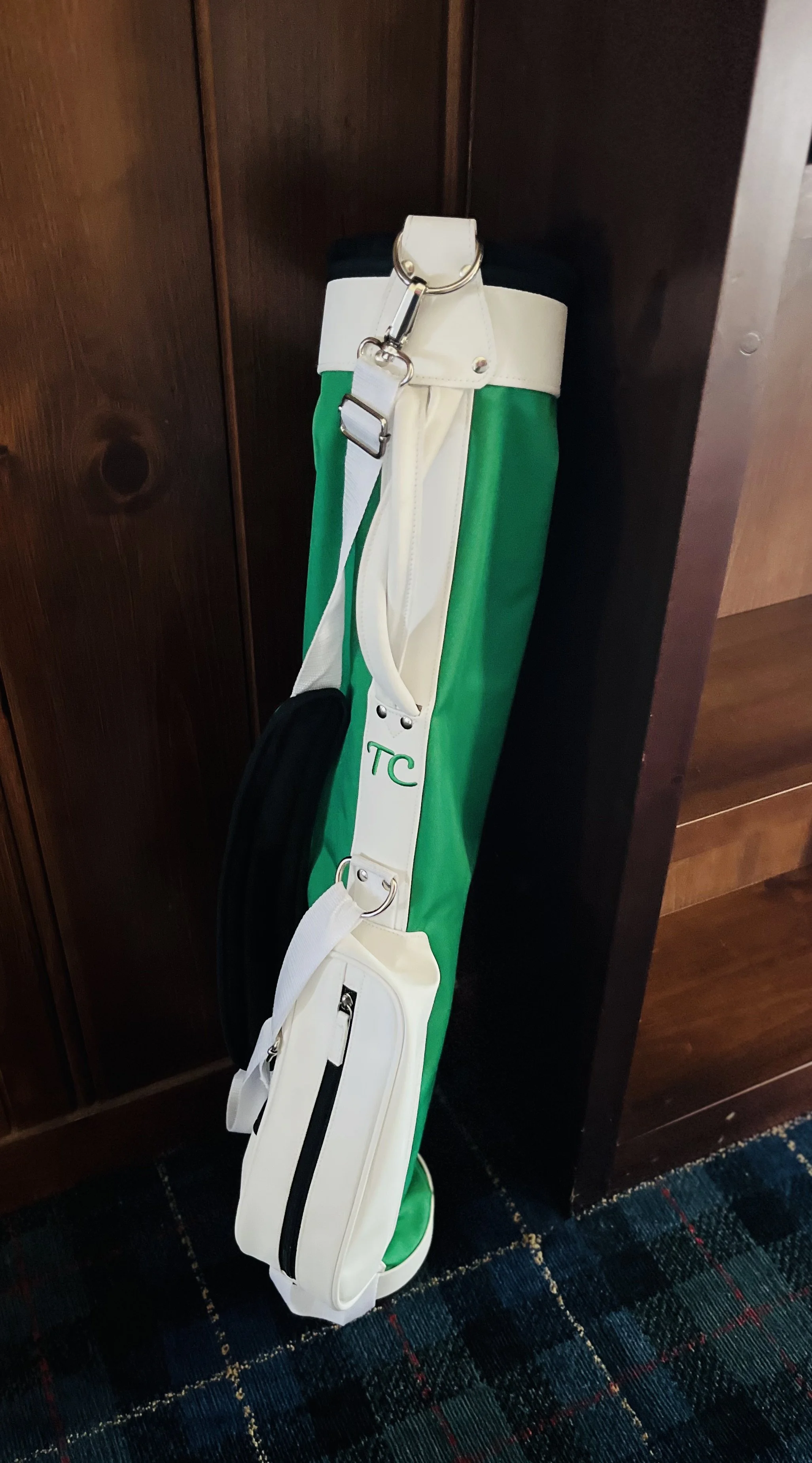 A golf bag with green, white, and black sections, standing upright next to a dark wooden cabinet on a patterned carpeted floor.