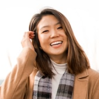 A smiling woman with shoulder-length hair wearing a tan coat and a plaid scarf.