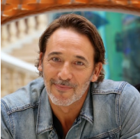 A man with dark brown hair, light beard, and mustache smiling, wearing a denim jacket, with a colorful amusement park background.
