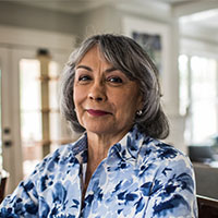 A woman with shoulder-length gray hair wearing a blue and white floral shirt standing in a well-lit room with large windows.