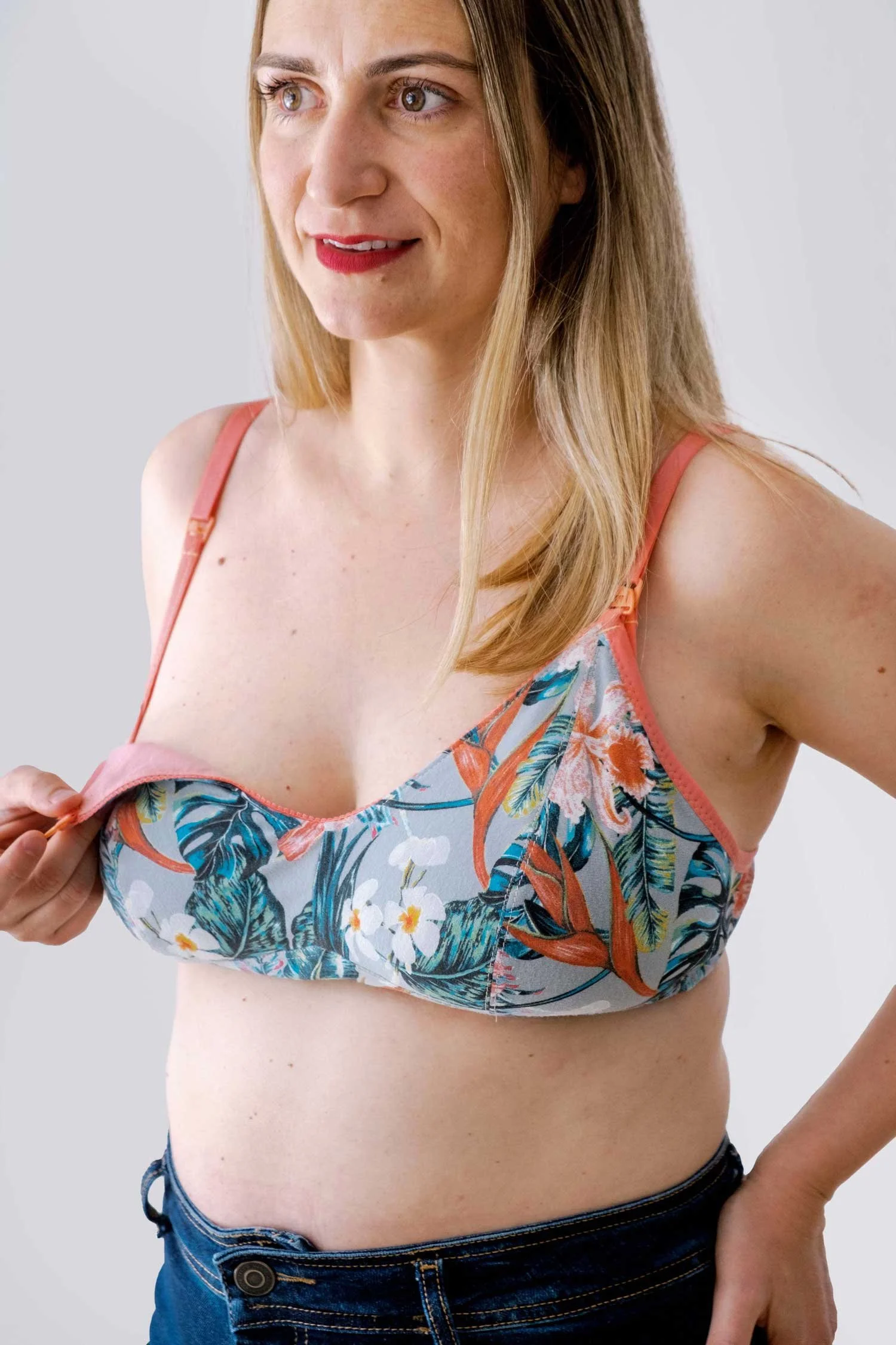 Mujer con cabello rubio, vestido de tirantes con estampado floral y azul, y pantalones vaqueros. Está en un fondo gris claro y se le ve la parte superior del cuerpo, levantando un poco la blusa.