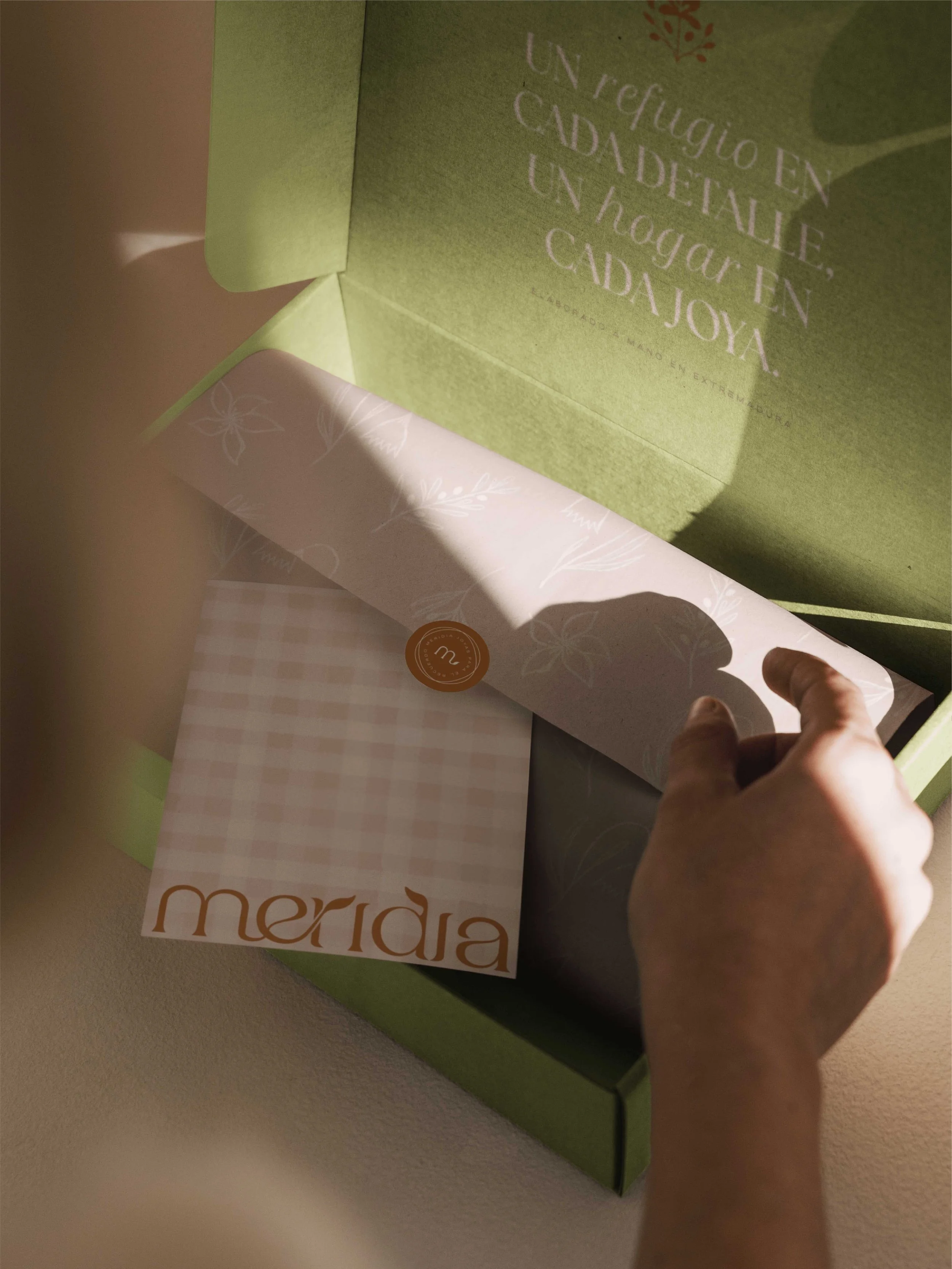 Caja de envío abierta en color verde oliva con papel de seda ilustrado y pegatina de marca creando un refugio visual.